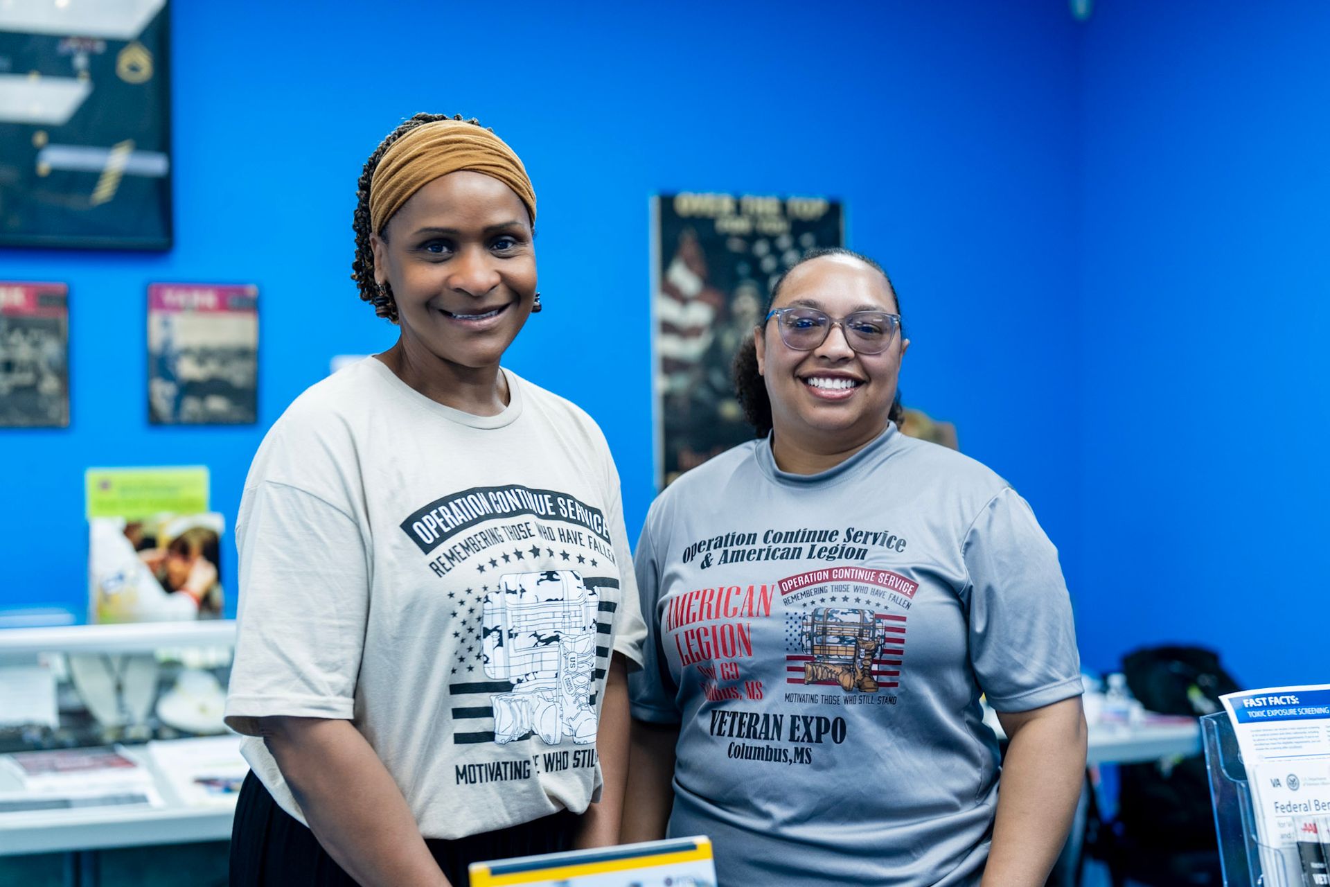 Two smiling people stand side-by-side in front of a bright blue wall with posters, wearing graphic t-shirts.