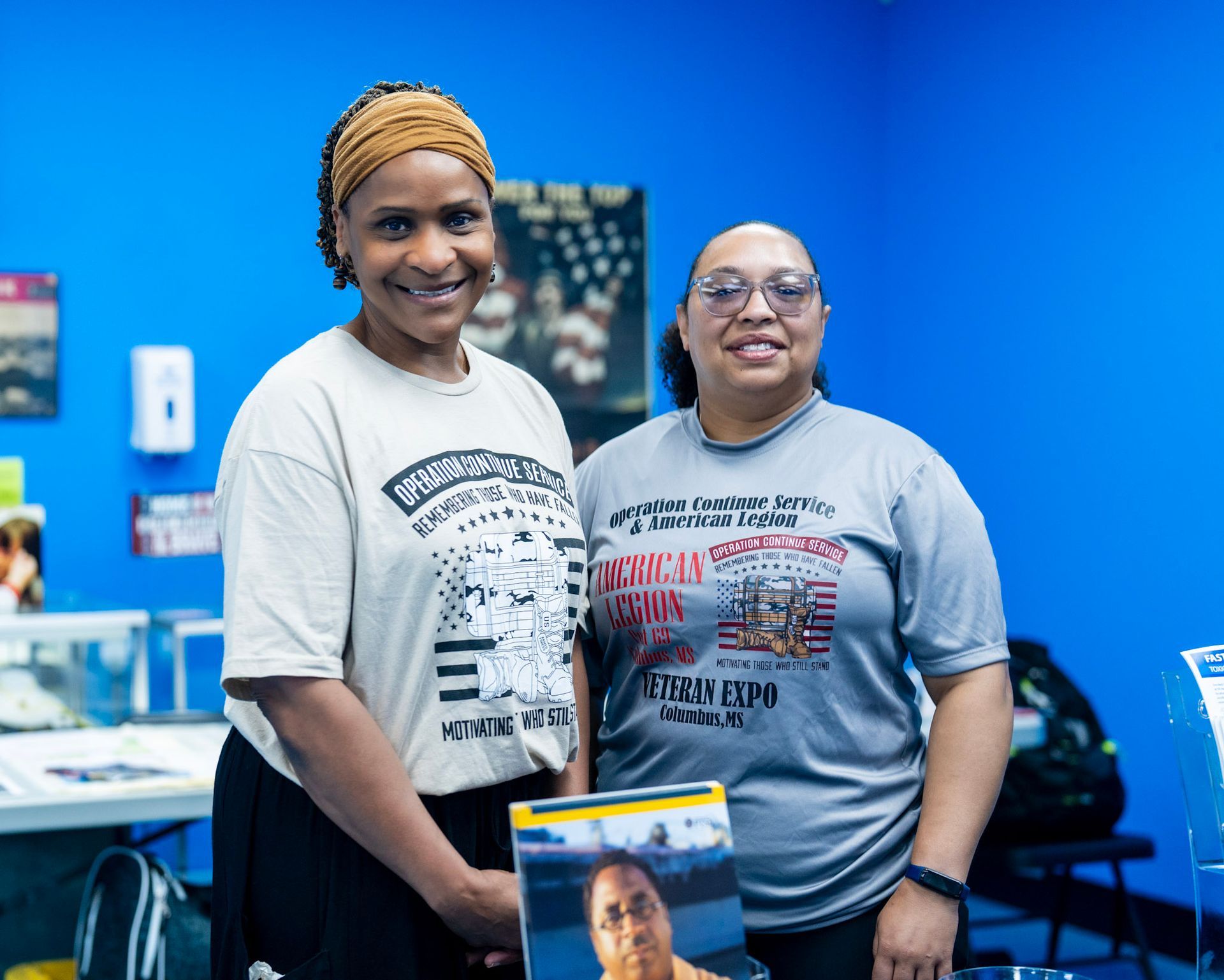 Two people smile while standing in front of a blue wall; one holds a framed photograph showing a person's headshot.