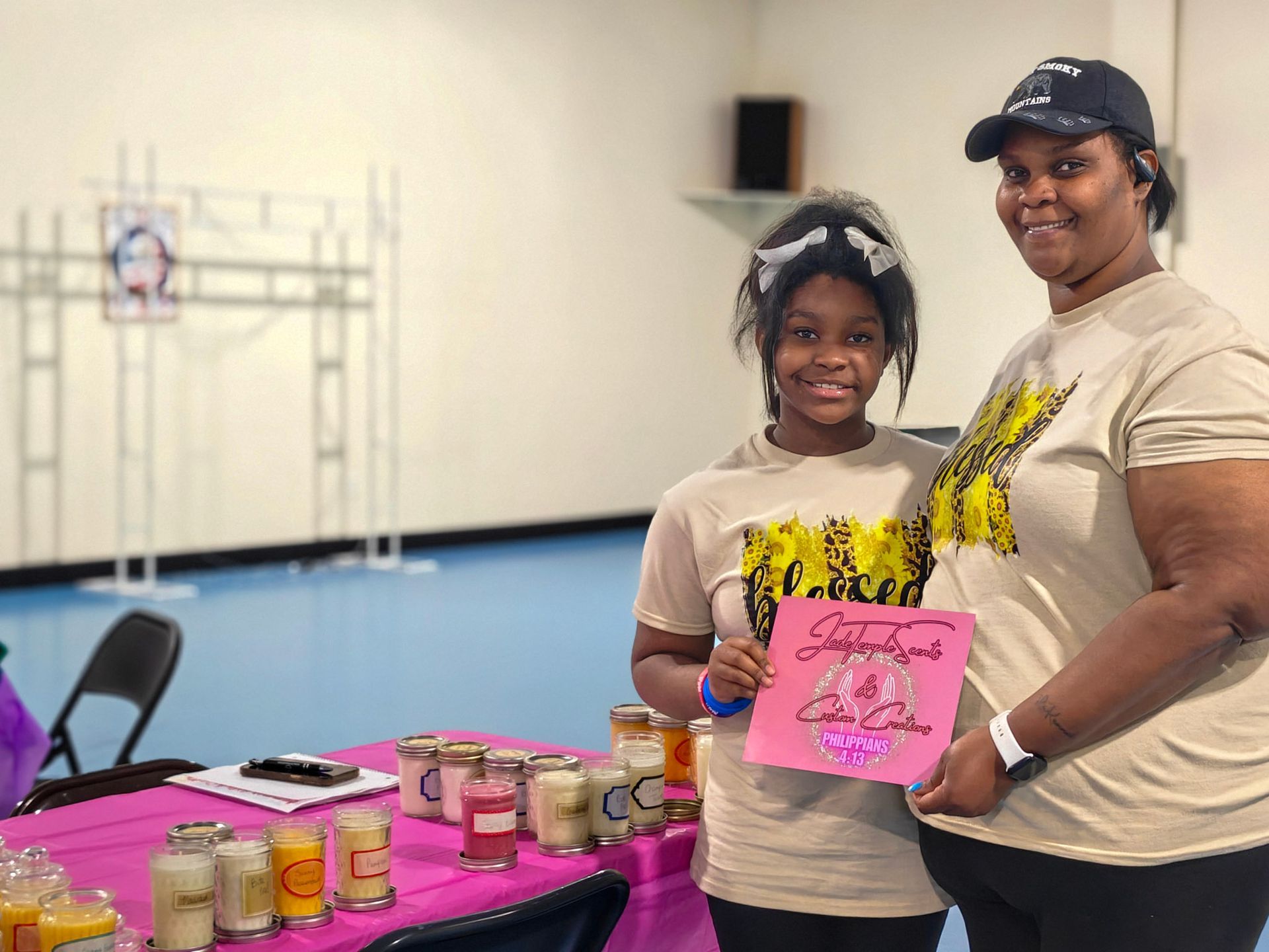 Two people standing behind a table displaying candles, holding a pink sign, in a room with a light blue floor.