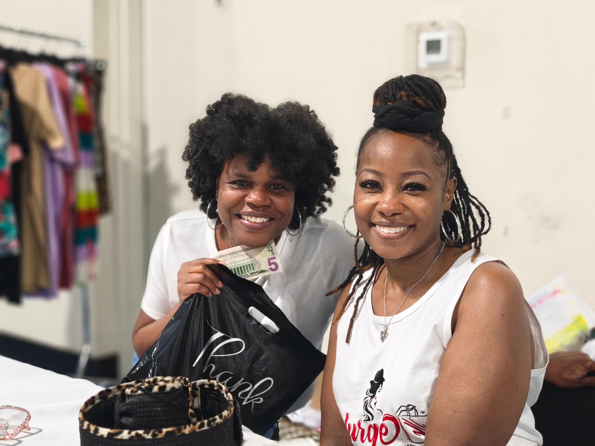 Two smiling people stand behind a table with a clothing rack in the background; one holds a black shopping bag.