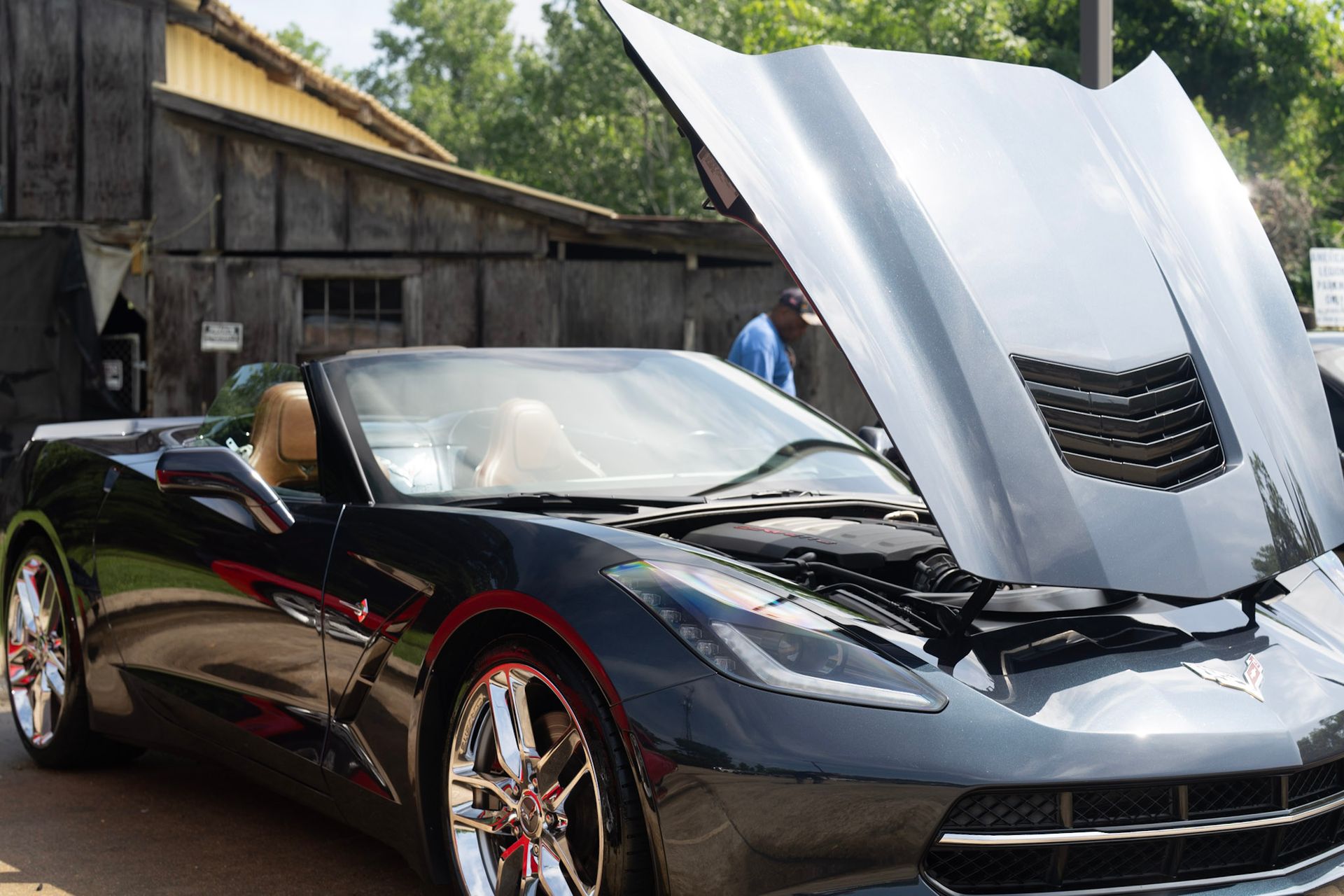 A black Chevrolet Corvette convertible with its hood open, parked in front of a rustic wooden building.
