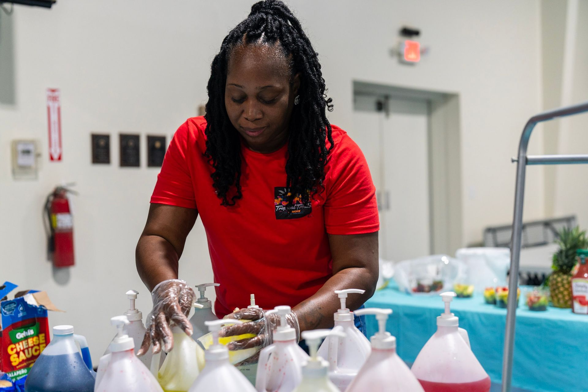 A person in a red shirt wears plastic gloves and prepares plastic squeeze bottles behind a table in a room.