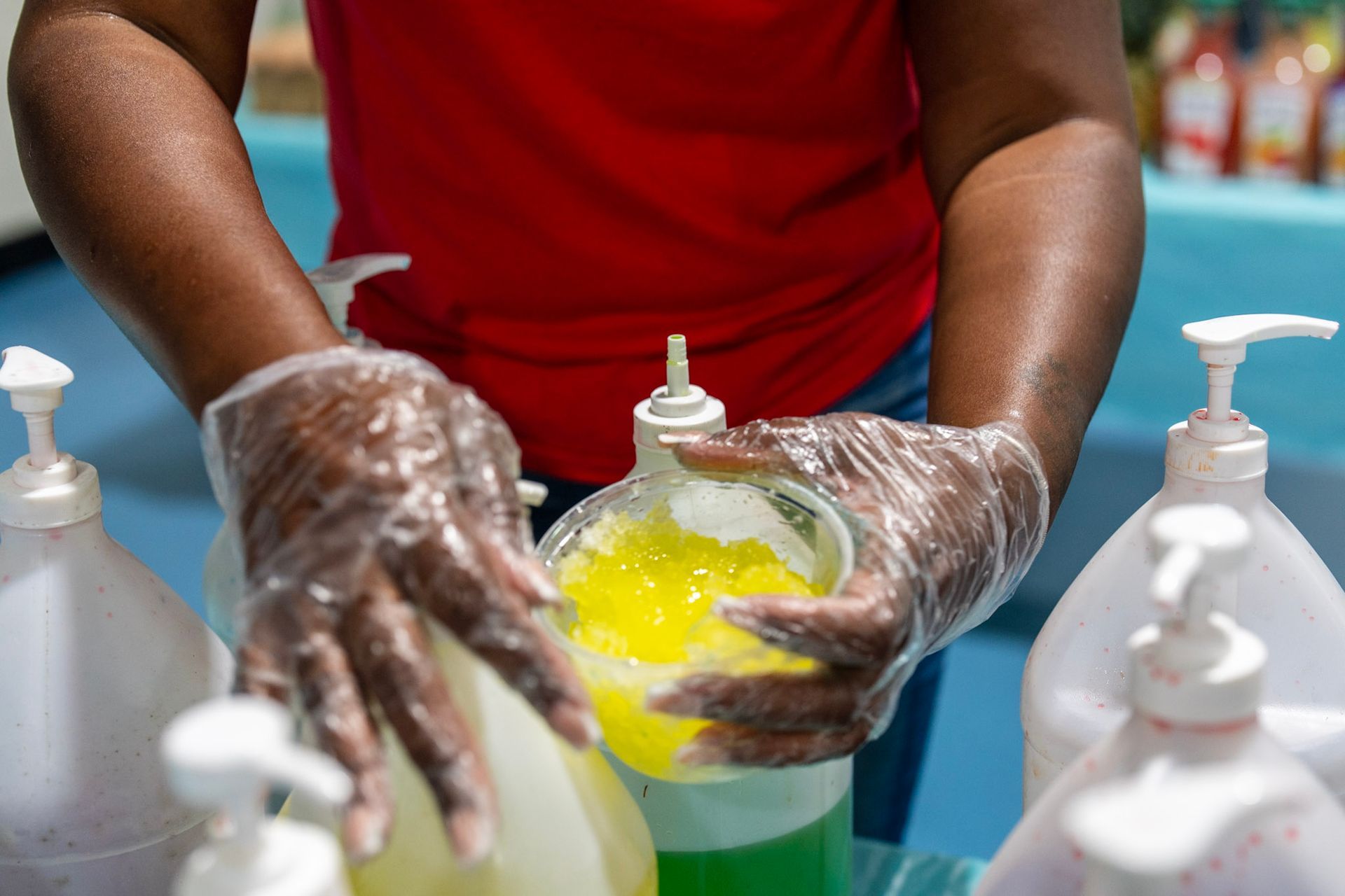 A person wearing clear plastic gloves handles a container of bright yellow gel surrounded by several pump bottles.