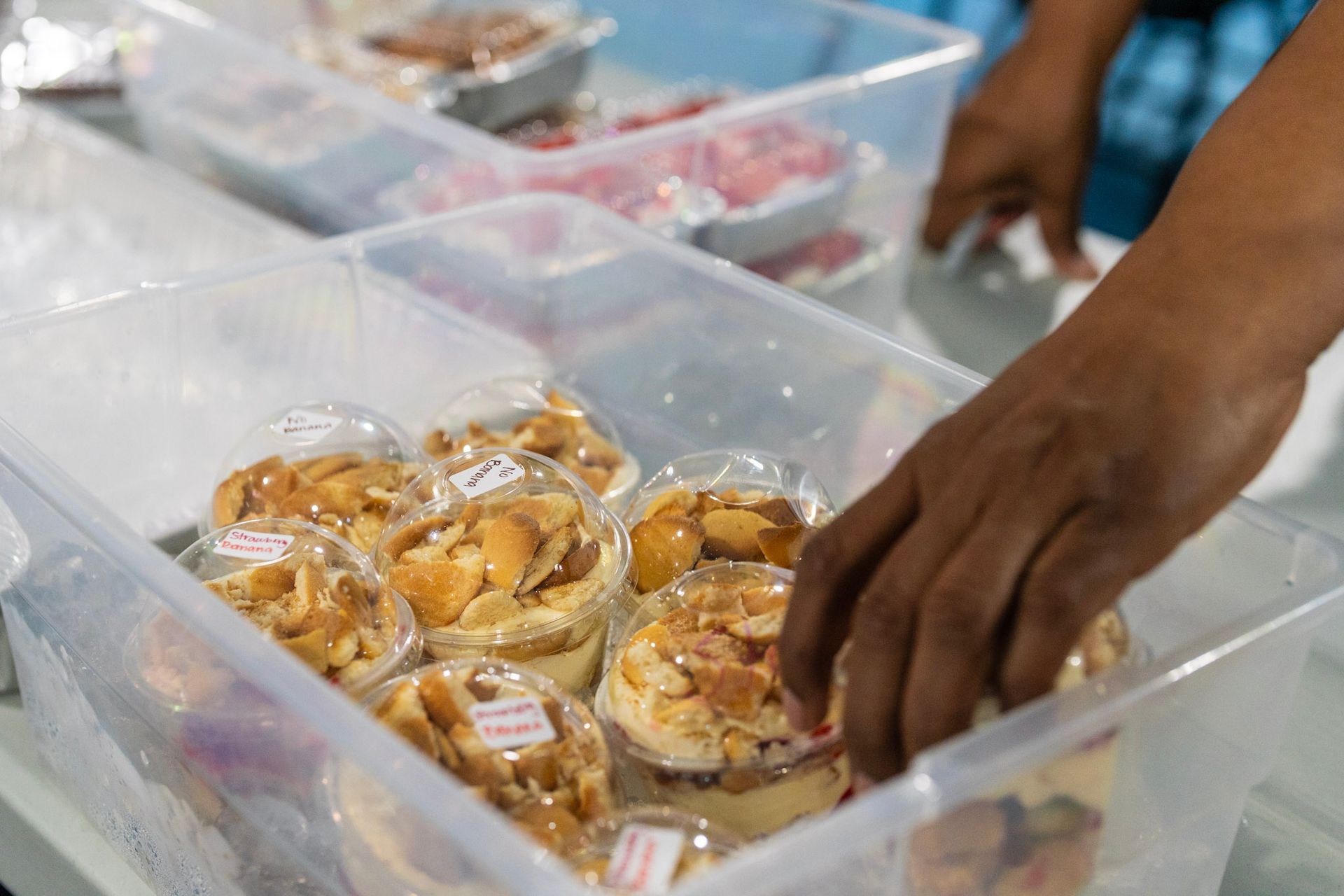 A hand reaching into a clear plastic bin filled with individual cups of banana pudding topped with vanilla wafers.