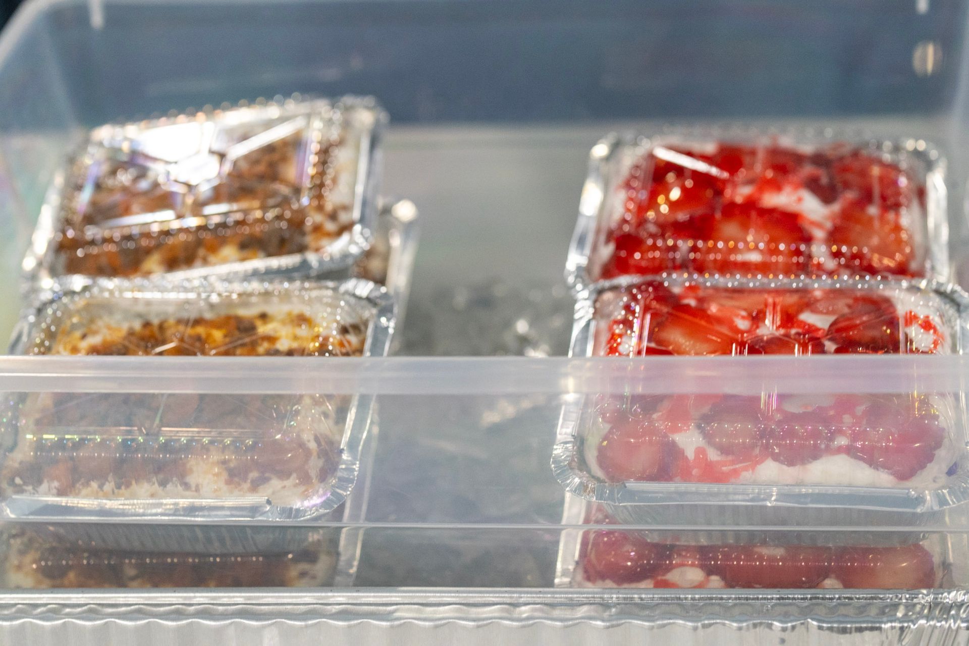 Foil-wrapped dessert portions, some topped with chocolate and others with glazed strawberries, inside a clear plastic bin.