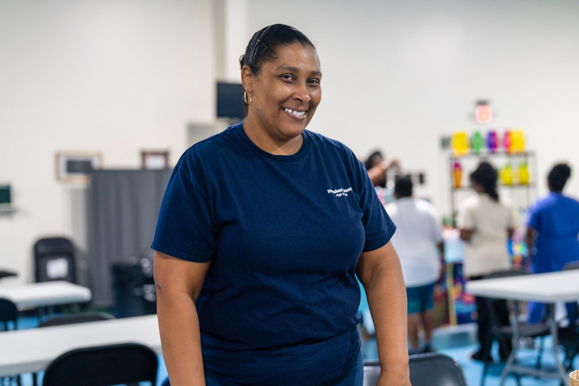 A smiling person in a dark blue shirt stands in a community space with tables and shelves in the background.