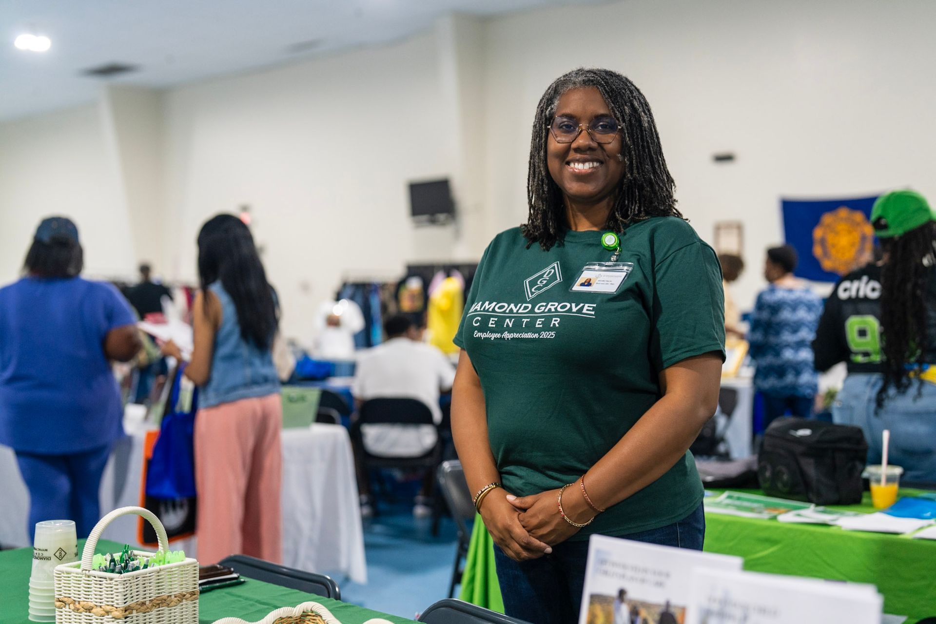 A smiling person wearing a green T-shirt stands in a bright room during an event, with other attendees in the background.