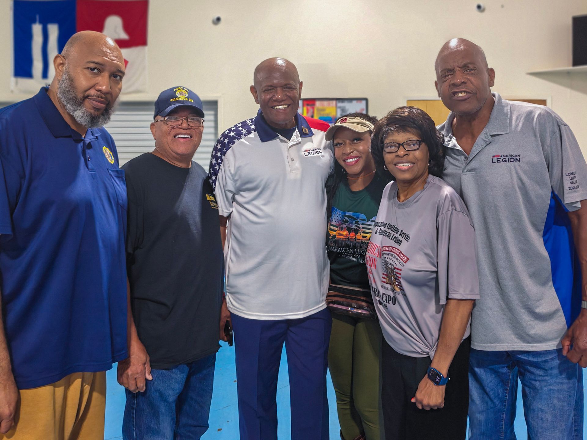 Six people stand together for a group photo indoors in front of a blue and white flag with a symbol.