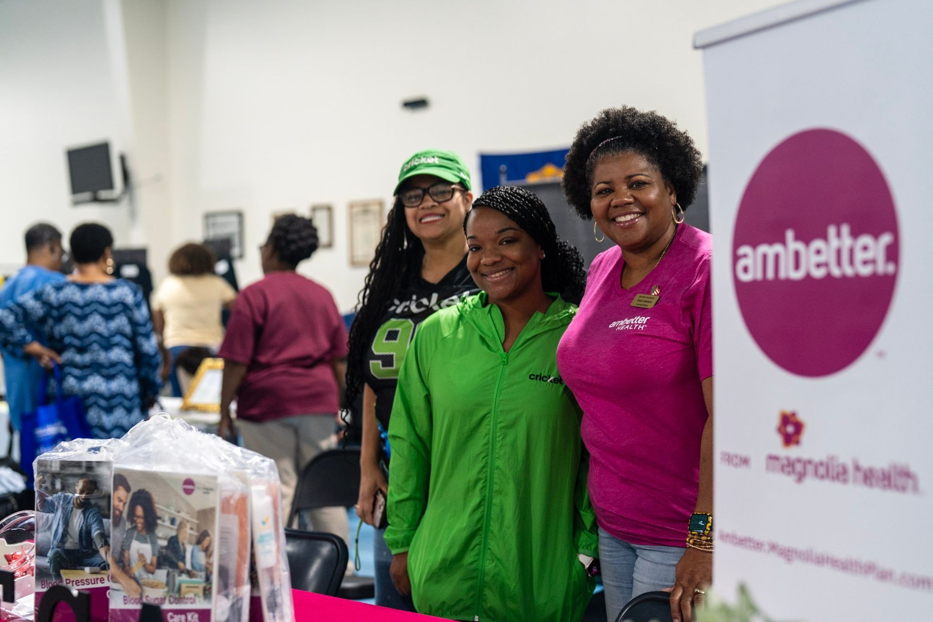 Three smiling people stand together behind an Ambetter from Magnolia Health banner at an indoor event.