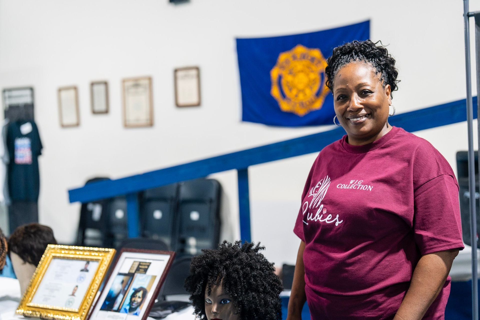A smiling person in a maroon t-shirt stands next to a mannequin head and framed certificates in an office or classroom.
