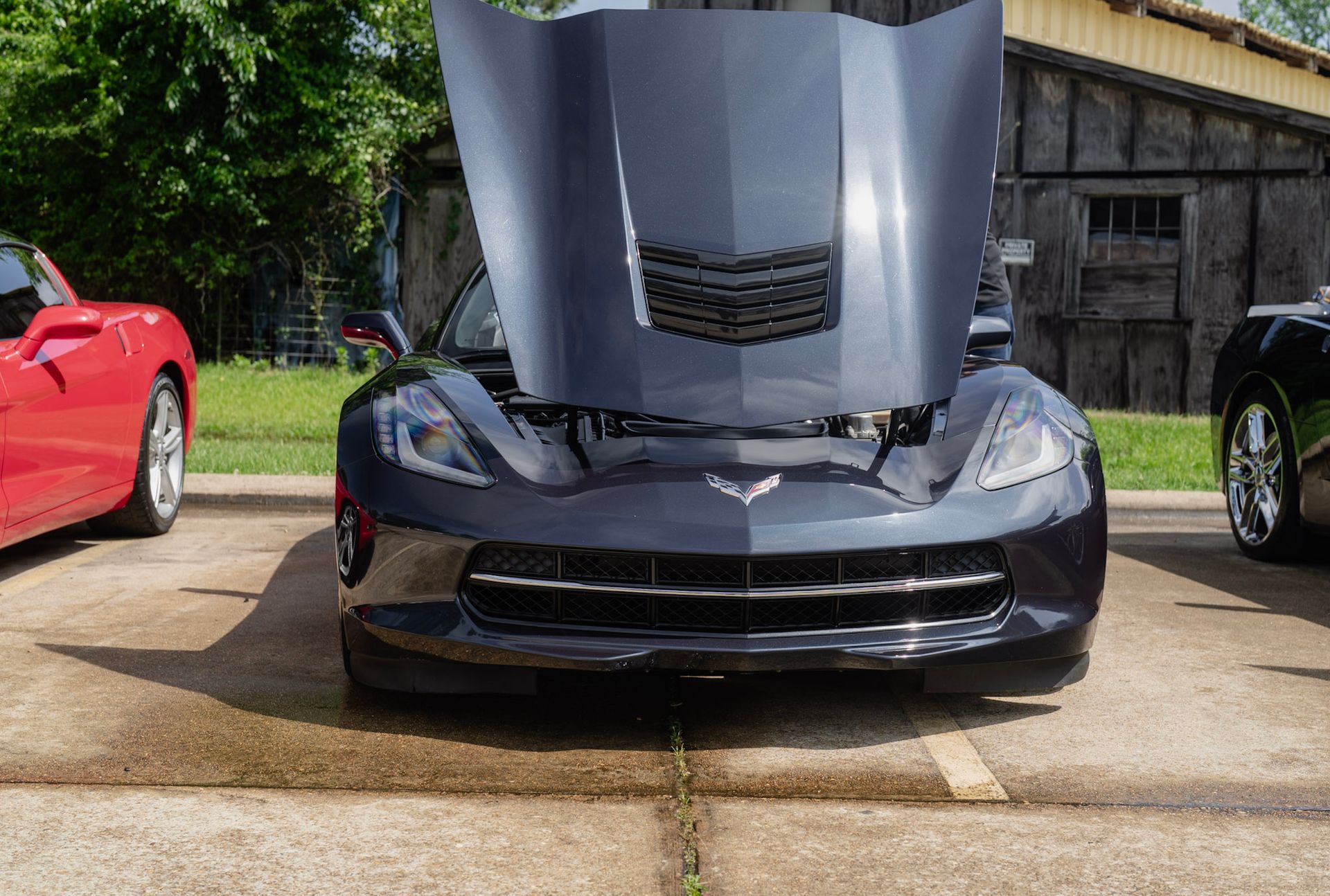 A dark gray Chevrolet Corvette parked outdoors with its hood open, flanked by a red car and a black car.