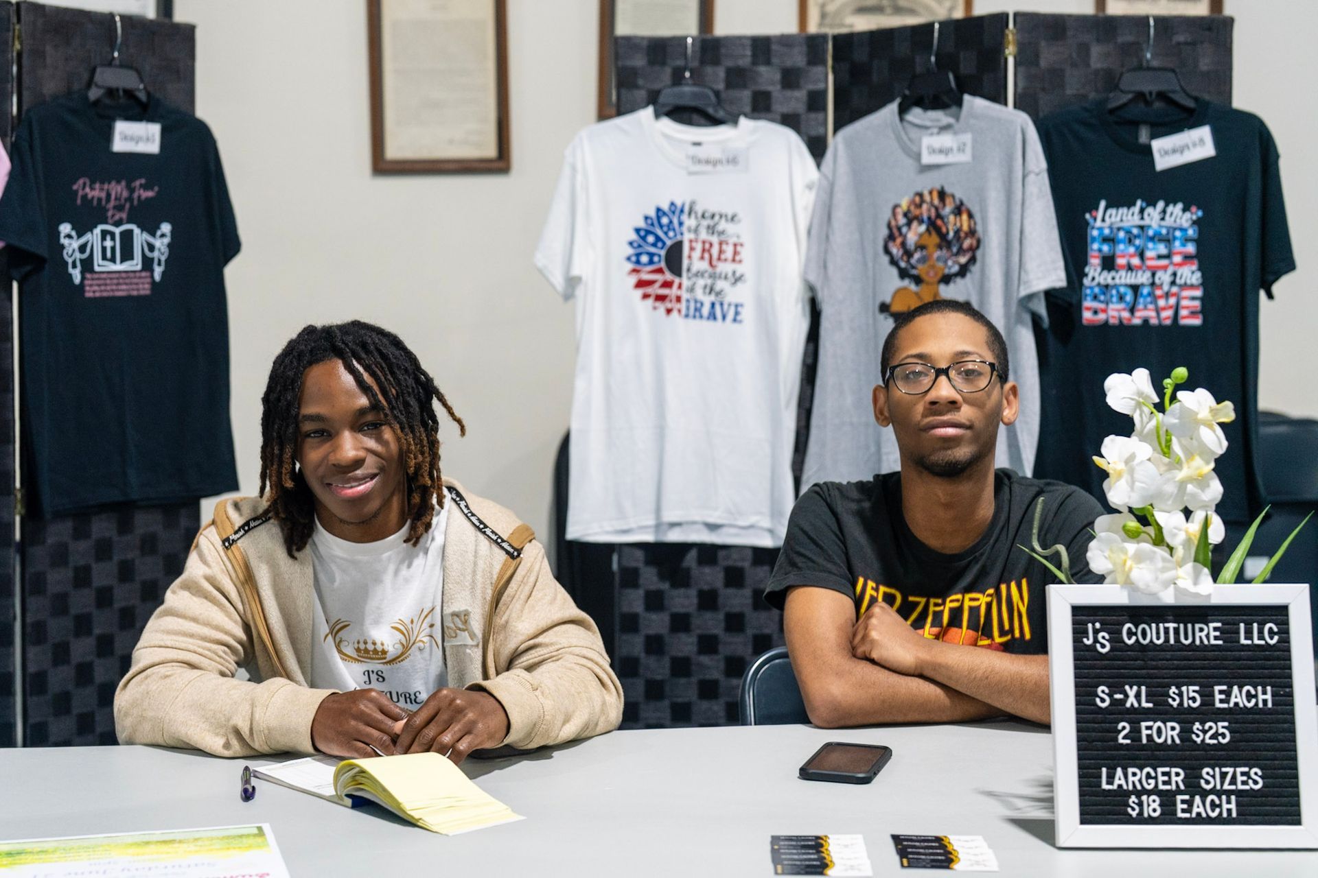 Two people sit at a table in front of display shirts, with a small sign listing prices for custom apparel.