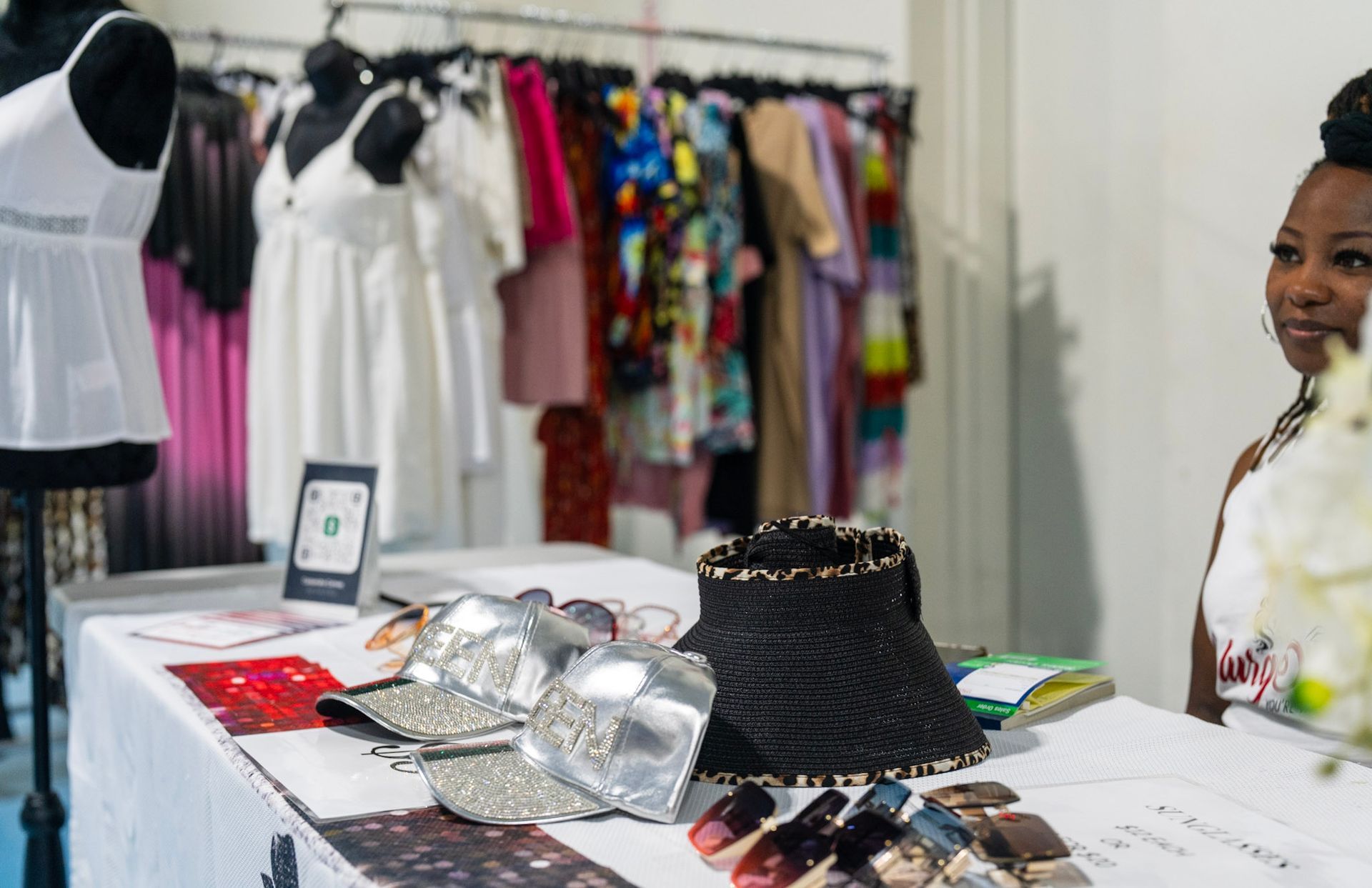 A shop table features sparkly silver accessories and a black hat, with clothing racks and a staff member in the background.