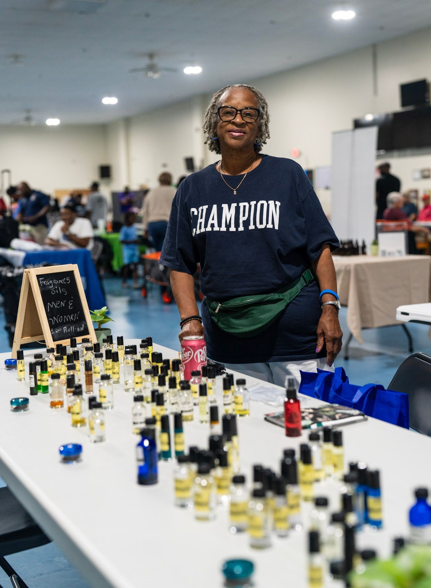 A person in a Champion shirt stands behind a table filled with small glass bottles at a community event.