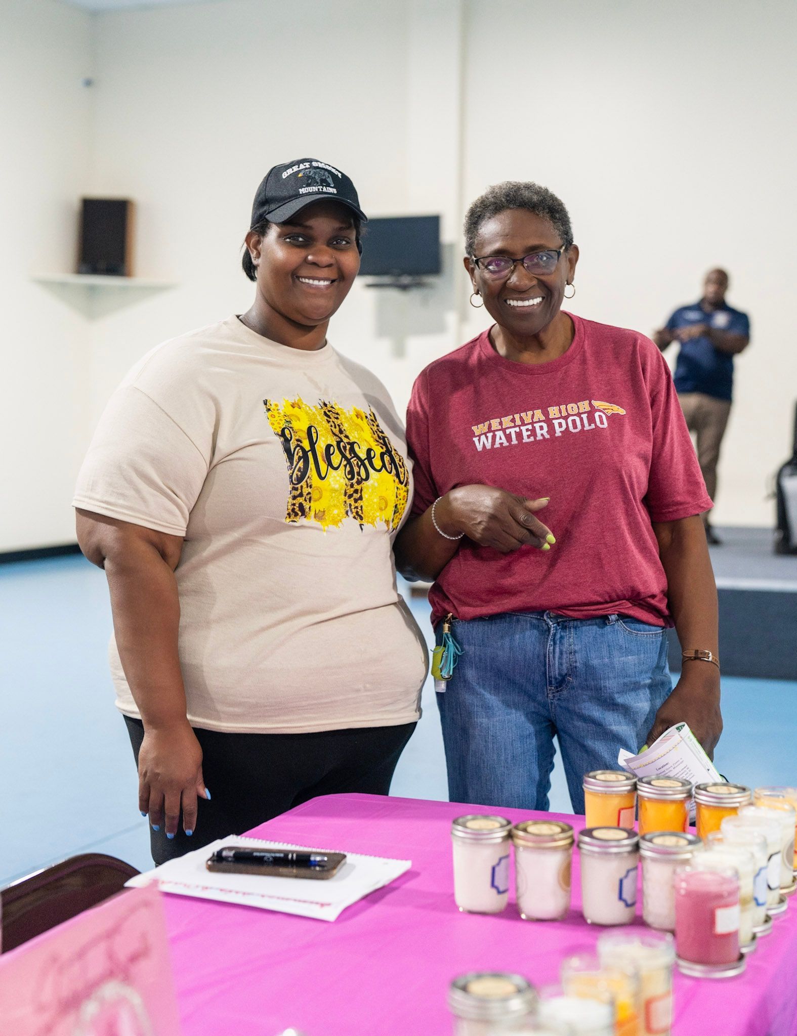 Two people smiling behind a pink-covered table displaying small candles in jars, with a person in the background.