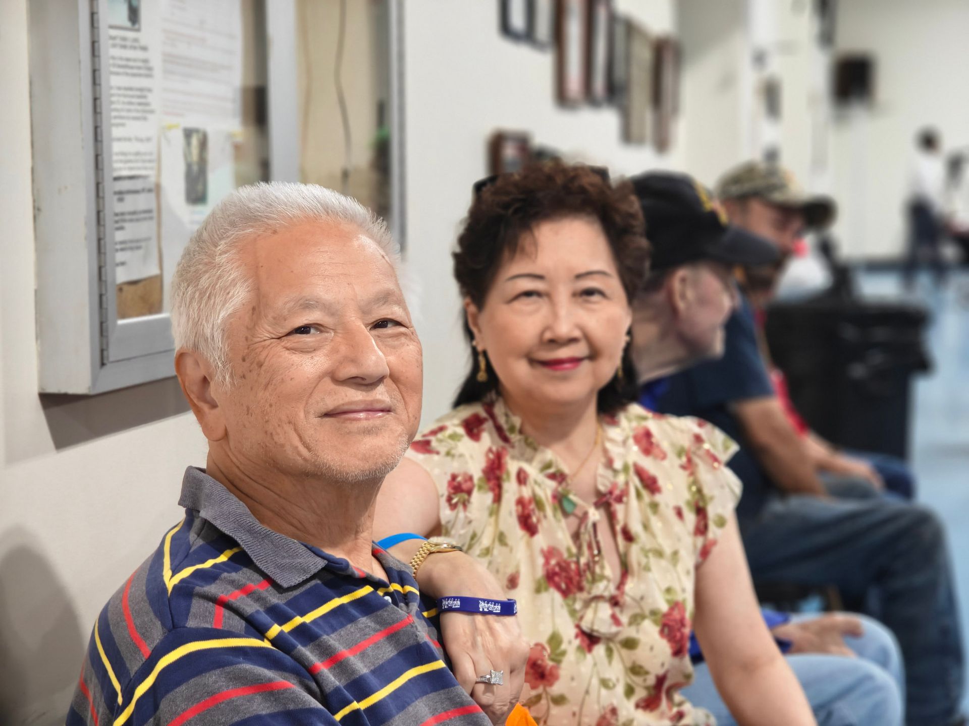A smiling man and woman sit side-by-side in an indoor public space with other people visible in the background.