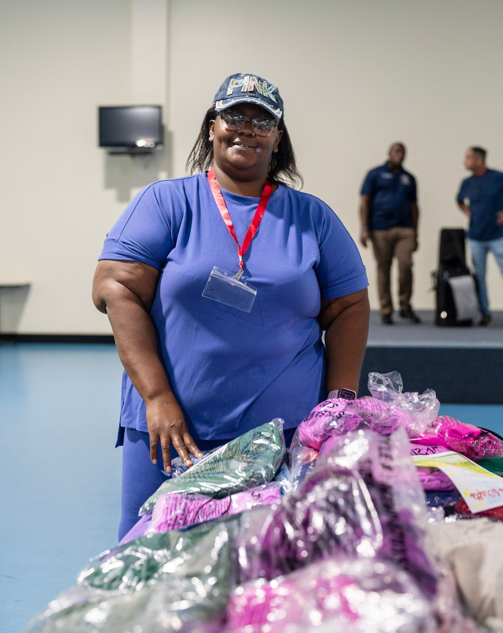 A smiling person in a blue shirt and hat stands behind a table filled with bagged items in a brightly lit indoor space.