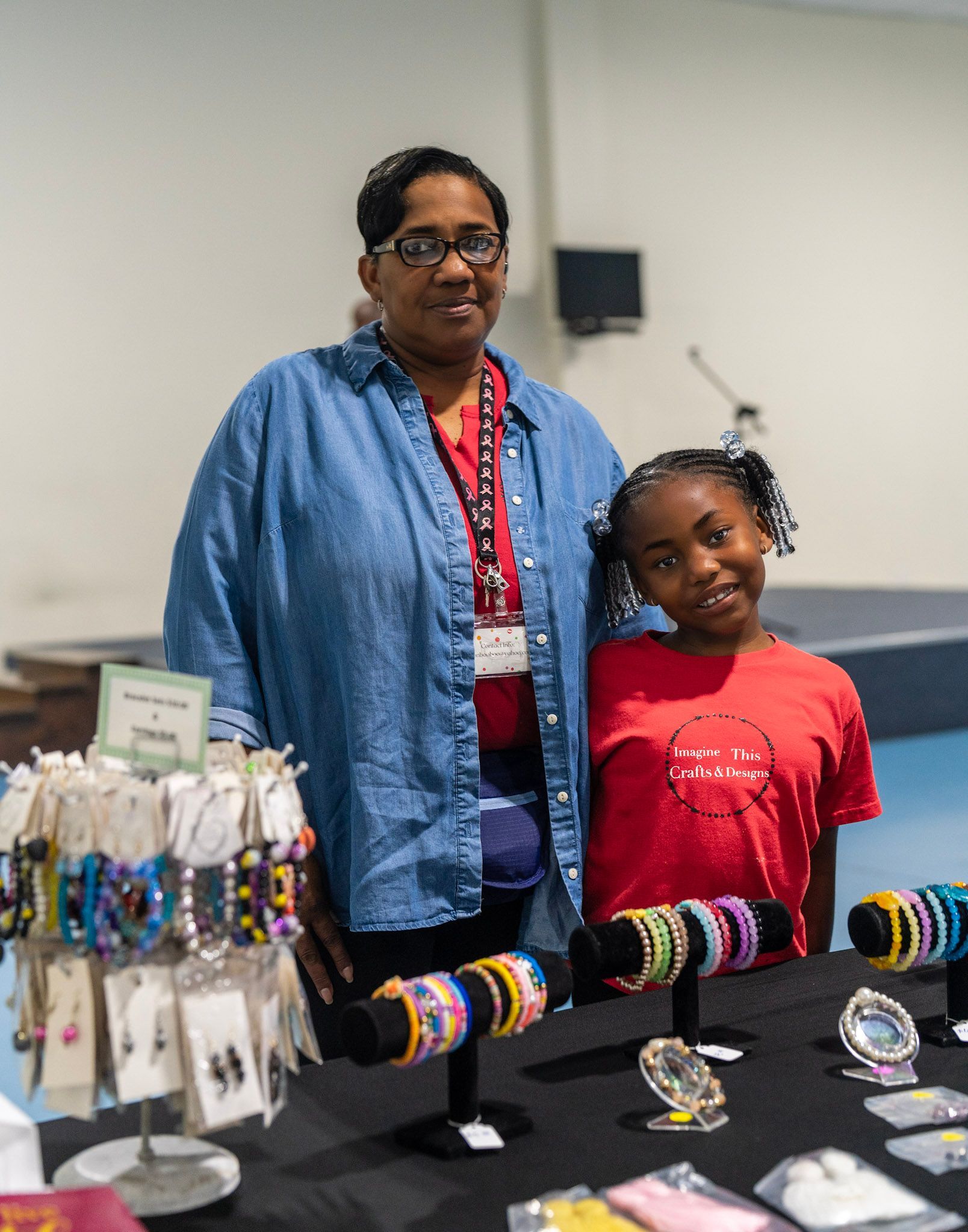 Two people stand behind a table display of colorful handmade bracelets, smiling at an event in an indoor space.