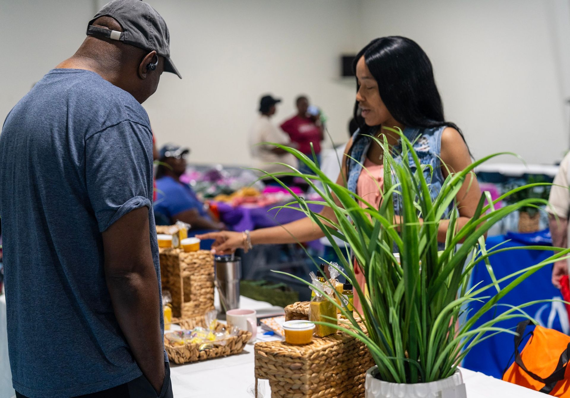 A person in a denim vest points to items on a vendor table while talking to another person at an indoor event.