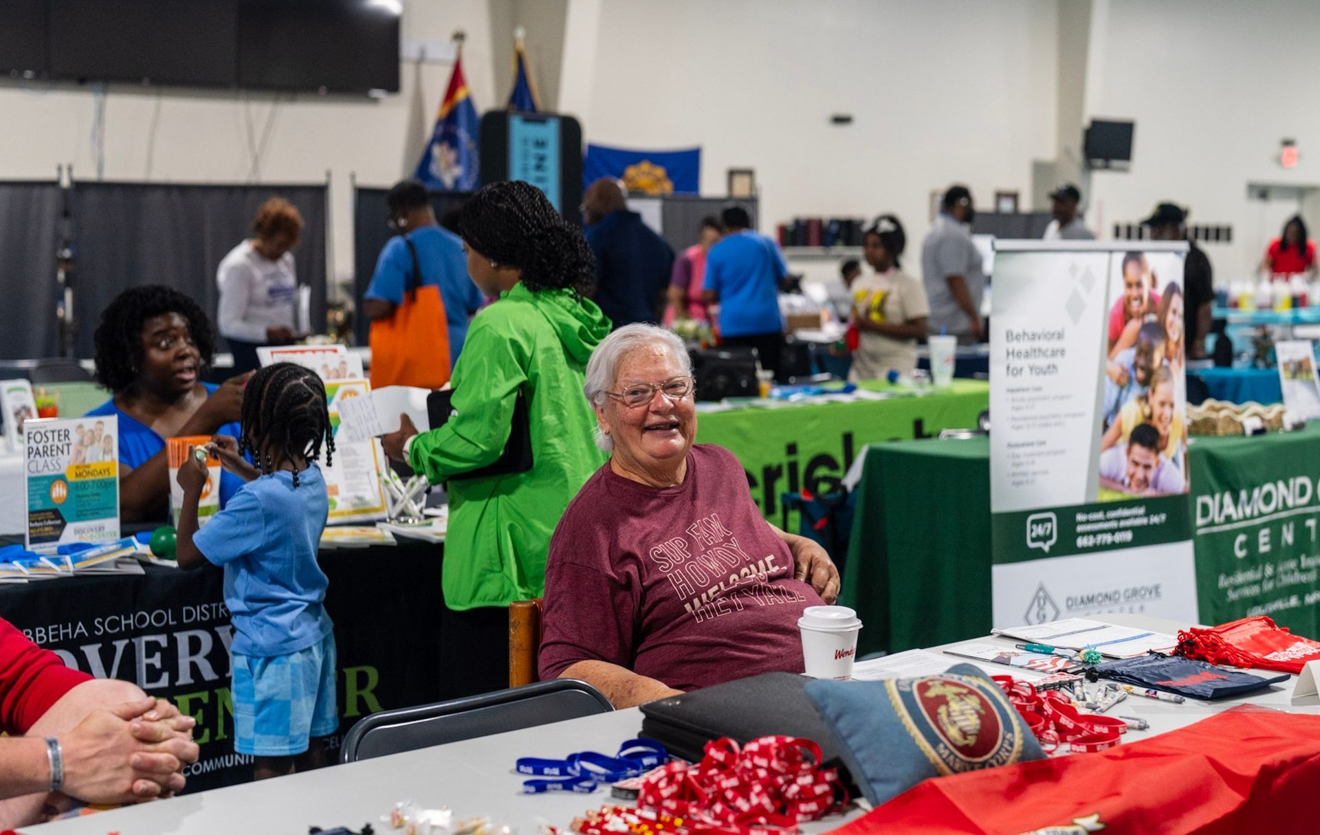 People gather at informational booths in an indoor community center, with a smiling individual at a table in the foreground.