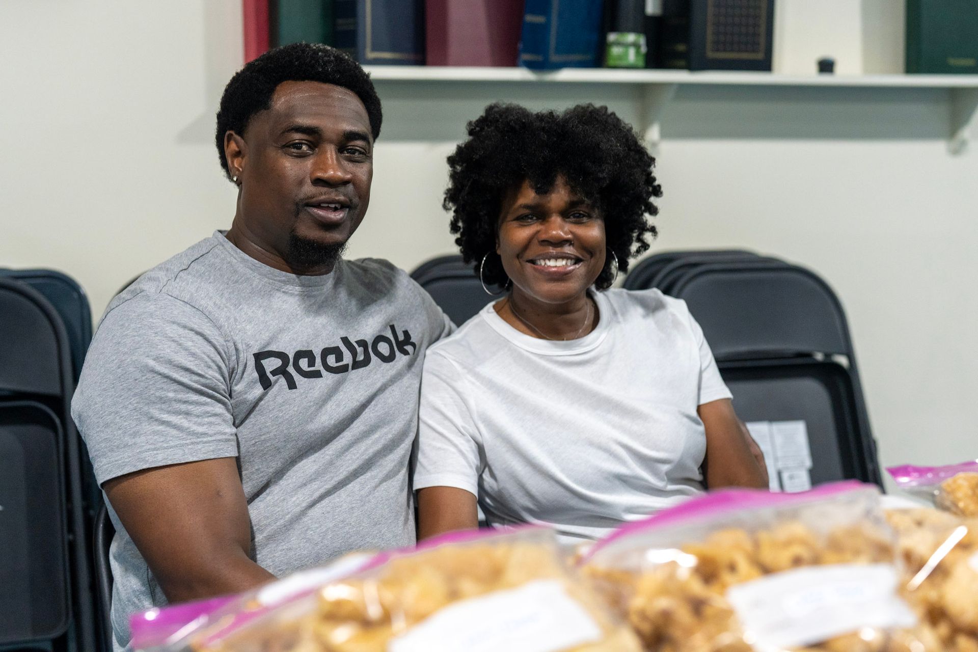 A man in a gray Reebok t-shirt and a woman in a white t-shirt sit together in front of plastic bags of food.