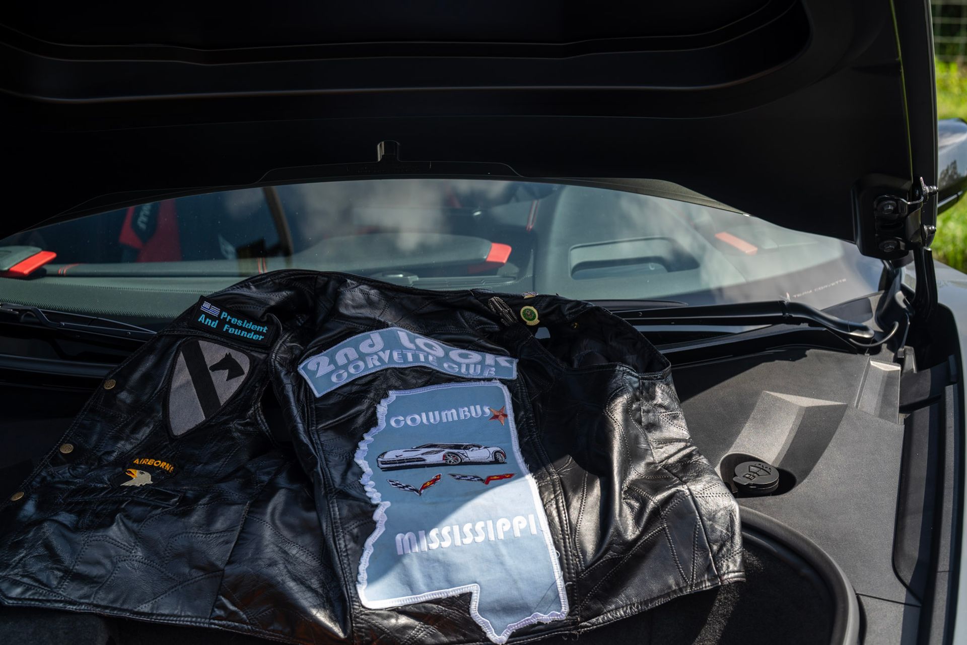 A black leather jacket with Mississippi and military-themed patches rests in the open front trunk of a car.