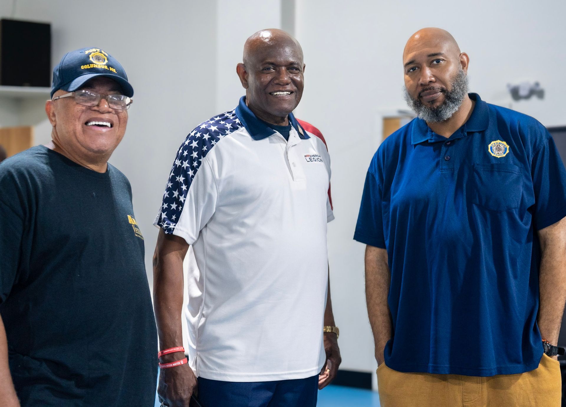 Three people stand together smiling, wearing casual shirts in an indoor setting.