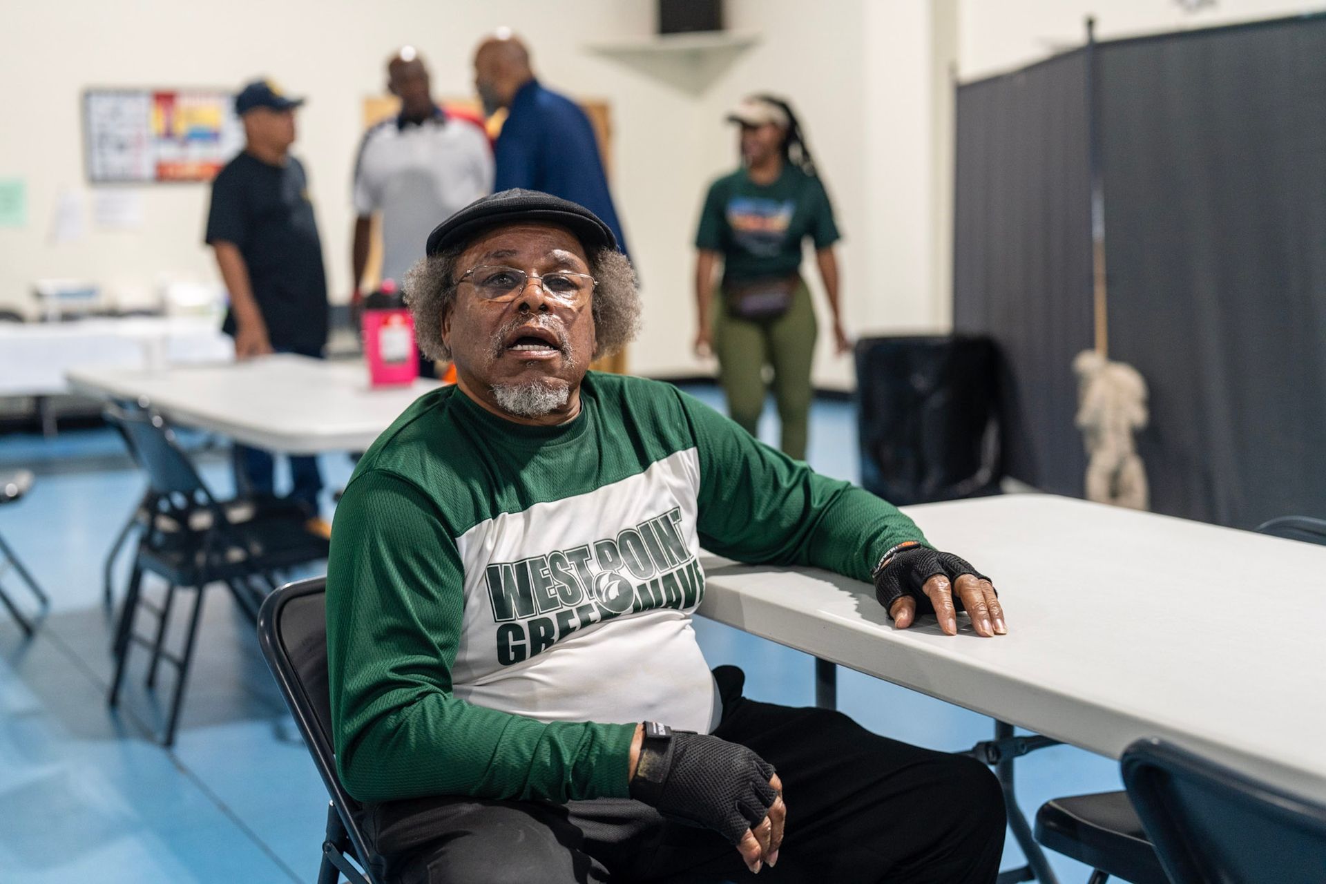 An older person in a green and white long-sleeved shirt sits at a table in a bright room with other people in the background.