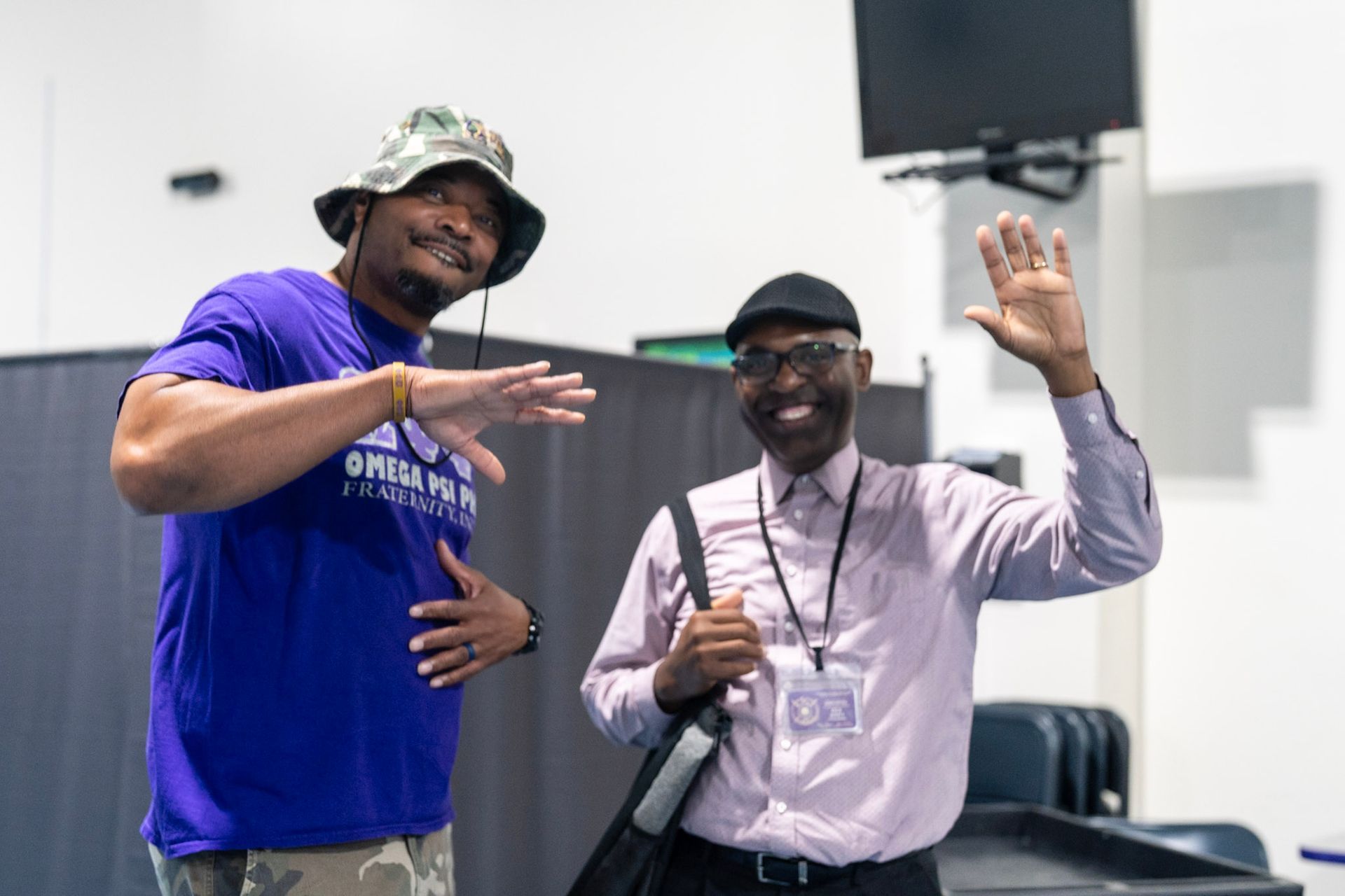 Two people smiling indoors, one wearing a purple t-shirt and bucket hat, the other wearing a dress shirt and waving.