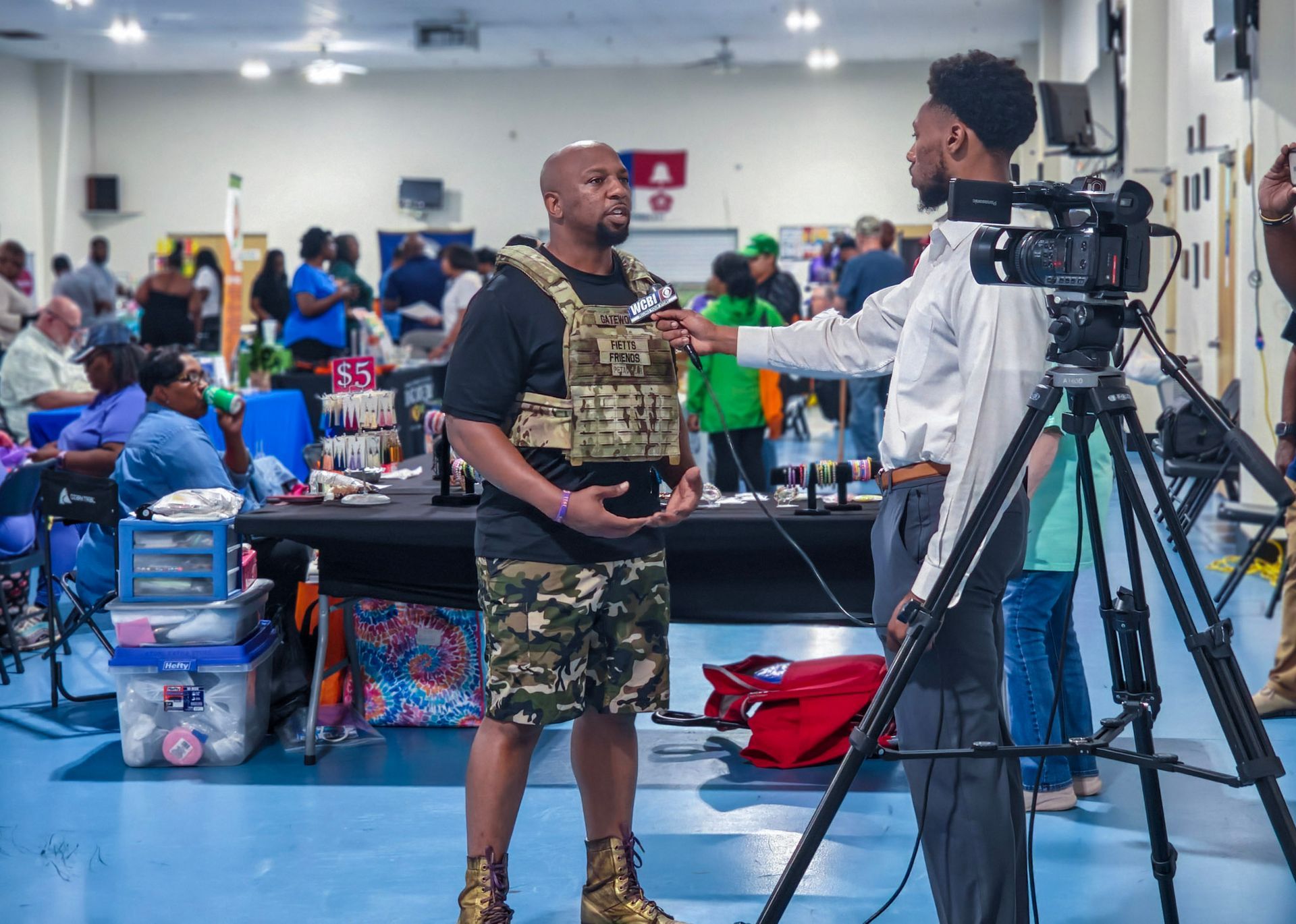 A reporter interviews a person wearing a tactical vest and camouflage shorts inside a community center.