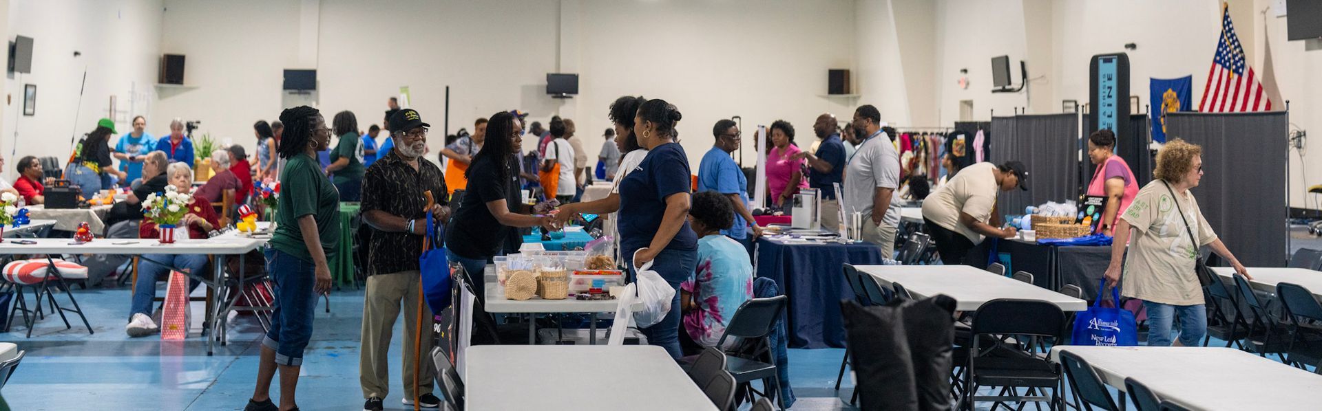 People gather in a large indoor event space with tables for a community meeting or resource fair.