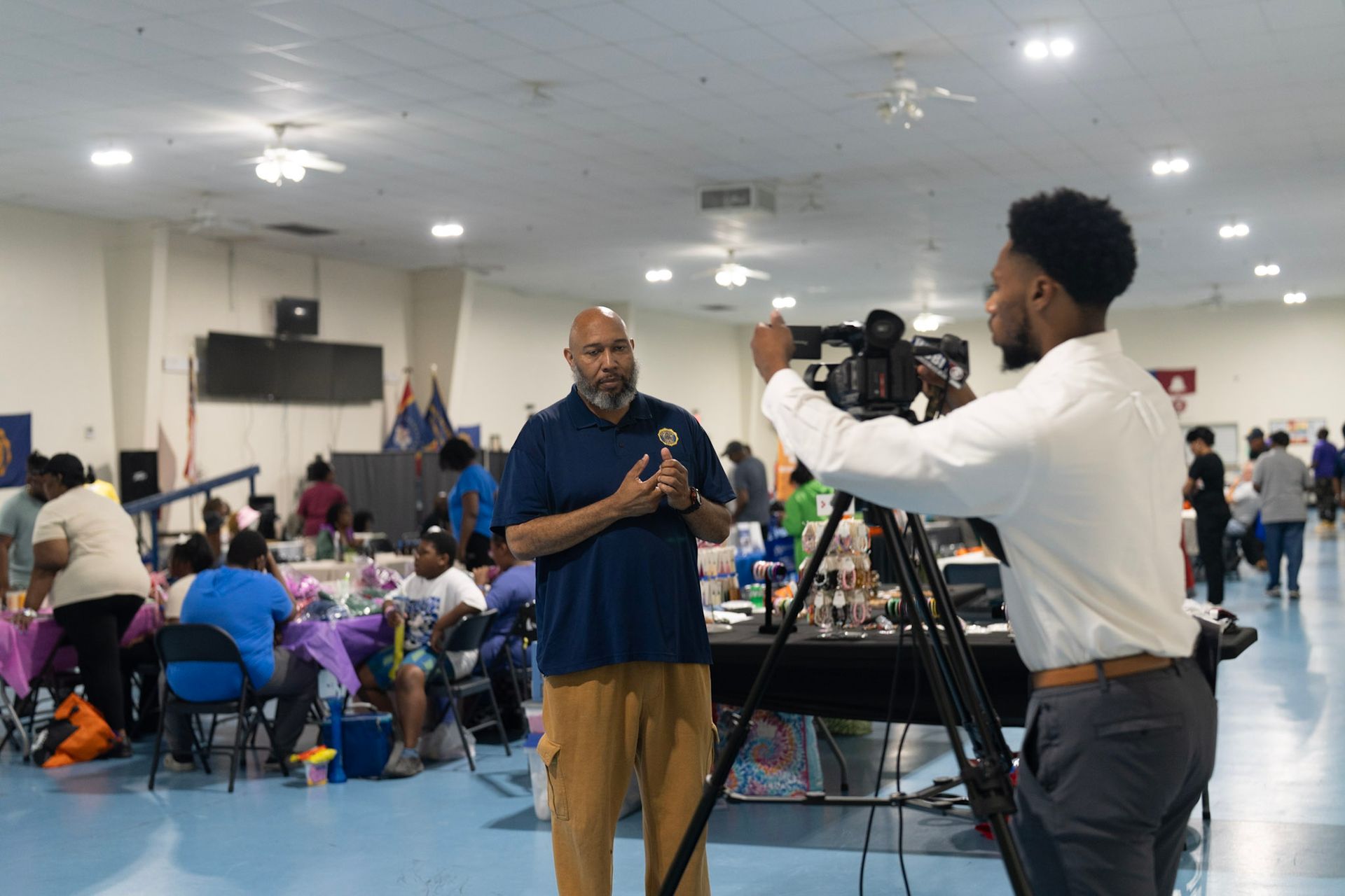 A person films an interview with a speaker in a community hall filled with people at tables.