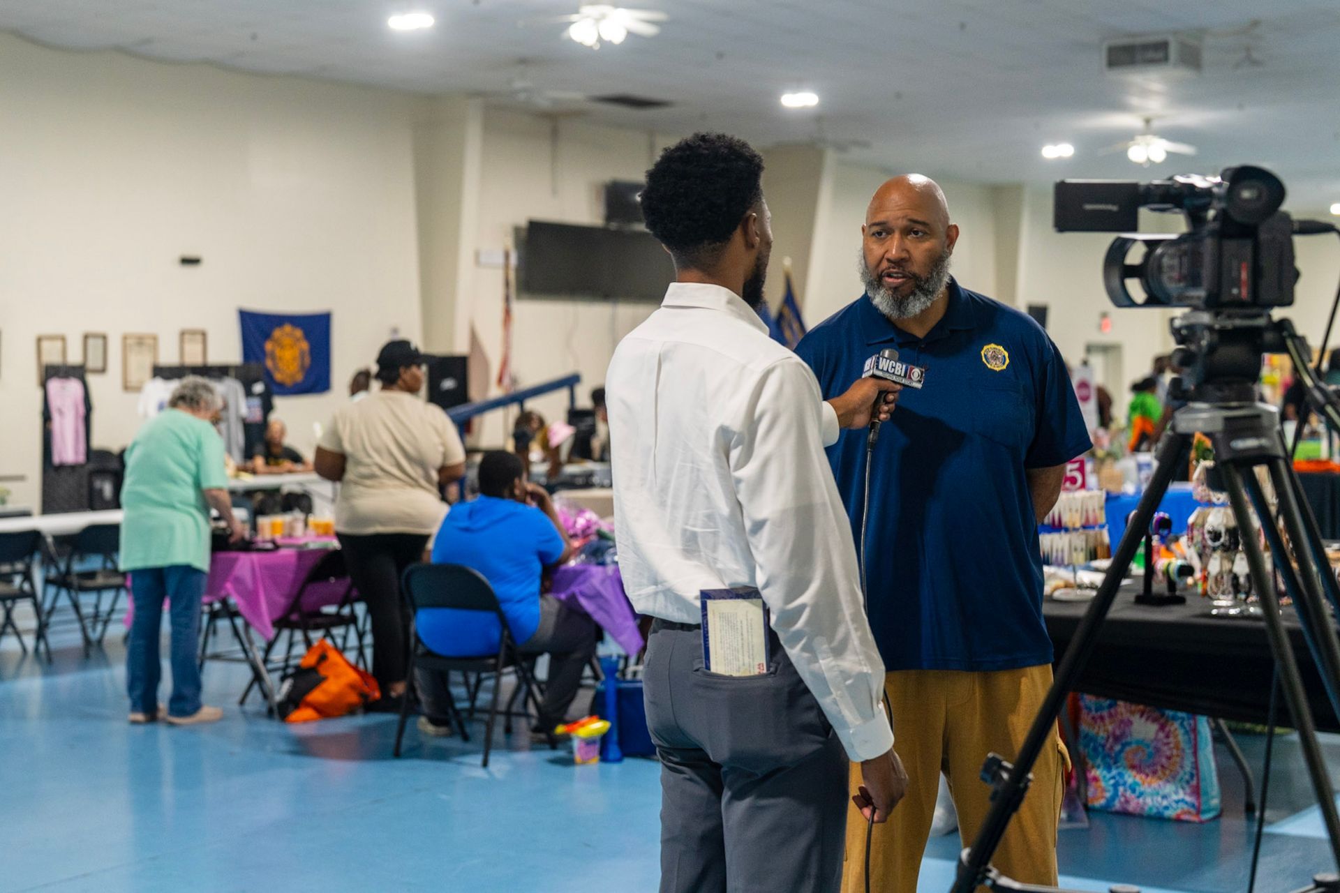 A reporter in a white shirt interviews a man in a navy polo shirt in a large, bright hall with tables and people.