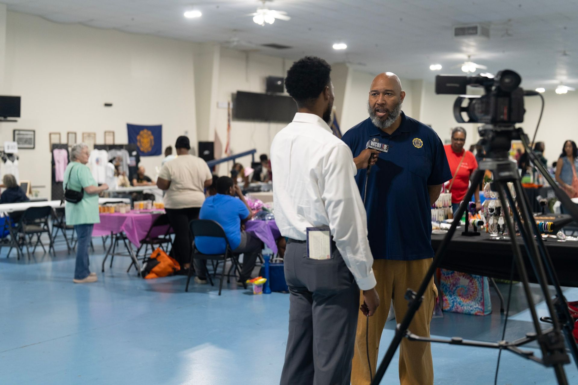 A reporter interviews a man inside a community hall, with a camera on a tripod in the foreground.