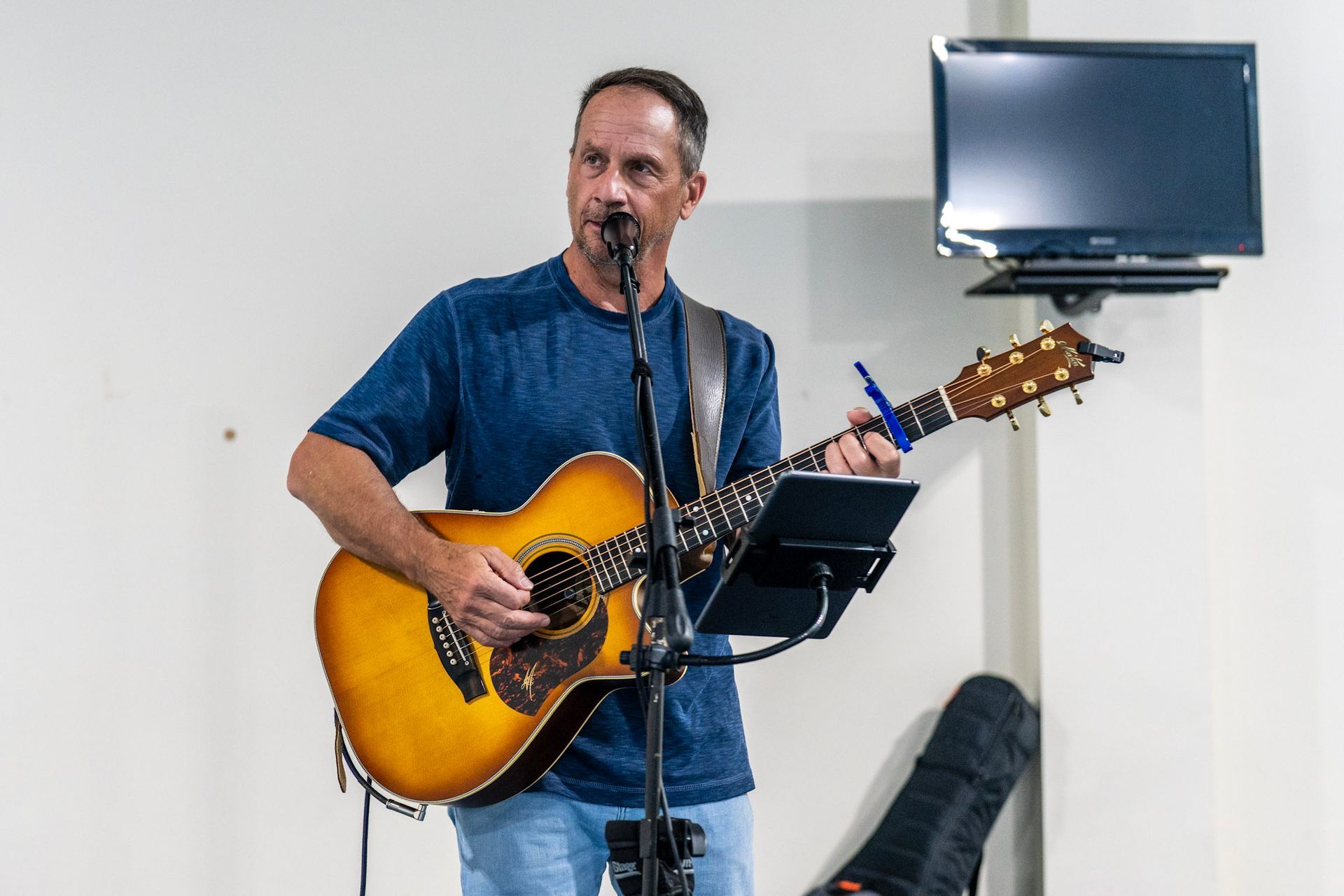 A musician plays an acoustic guitar and sings into a microphone while reading lyrics from a stand in a white room.