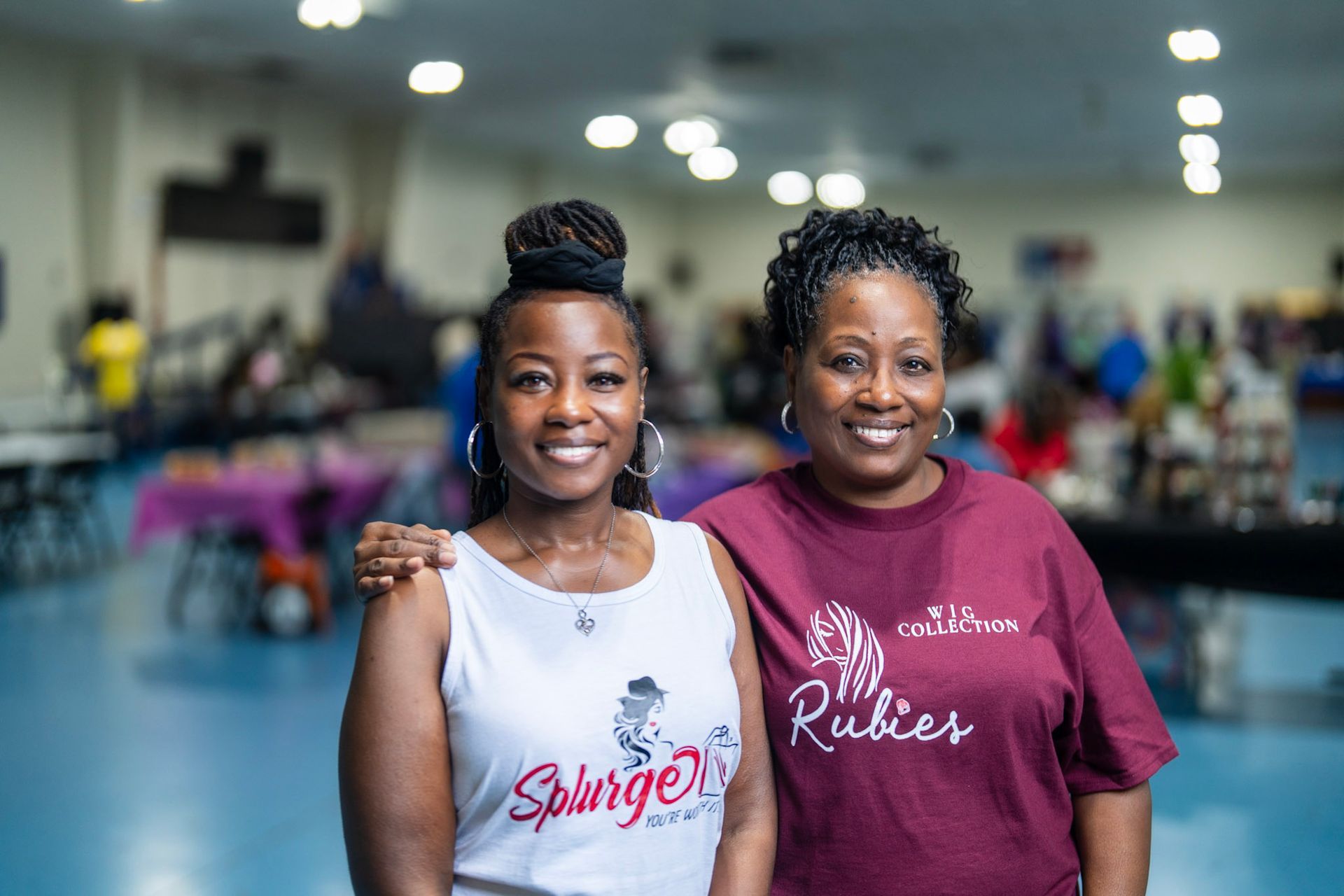 Two smiling people stand side-by-side in a large, open event space with tables in the background.