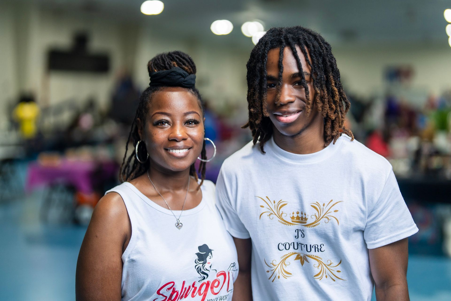 A smiling pair stands side-by-side in a large, blurred indoor event space, both wearing matching white graphic T-shirts.