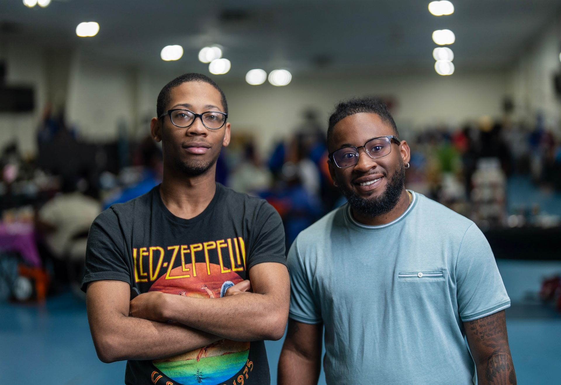 Two men pose for a portrait indoors, one in a Led Zeppelin t-shirt with arms crossed, the other in a light blue t-shirt.