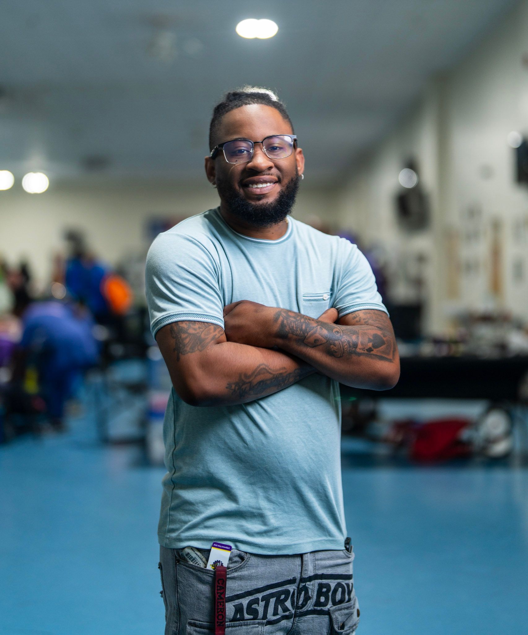 A smiling person wearing glasses and a light blue t-shirt stands with arms crossed in a gym setting.