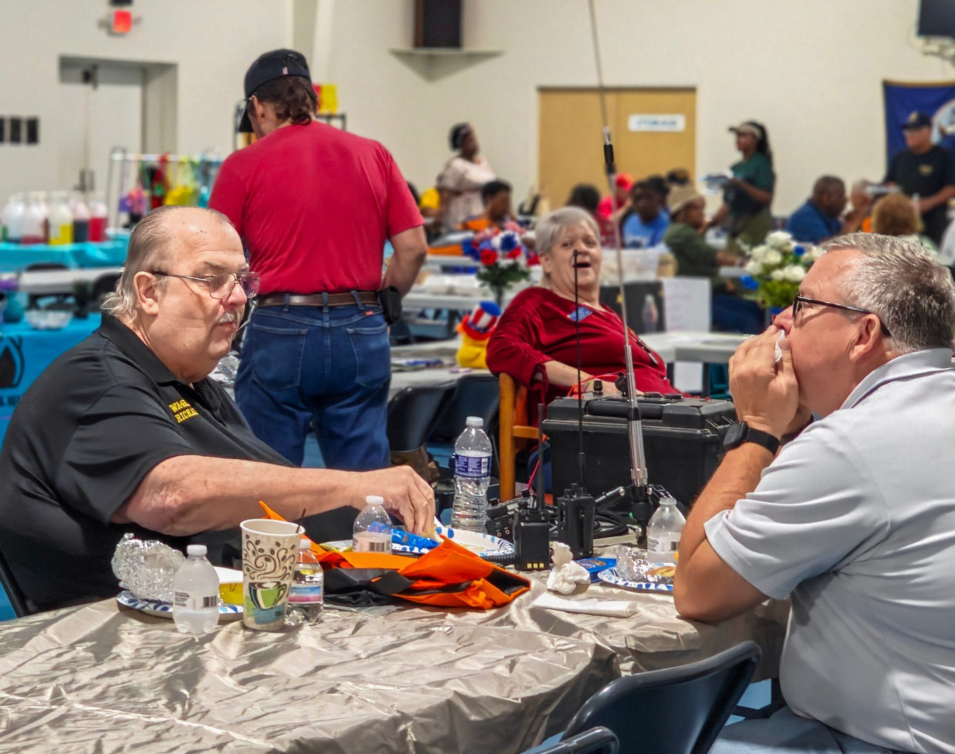 Two people sit at a table in a community hall, with others in the background, discussing items spread across the table.