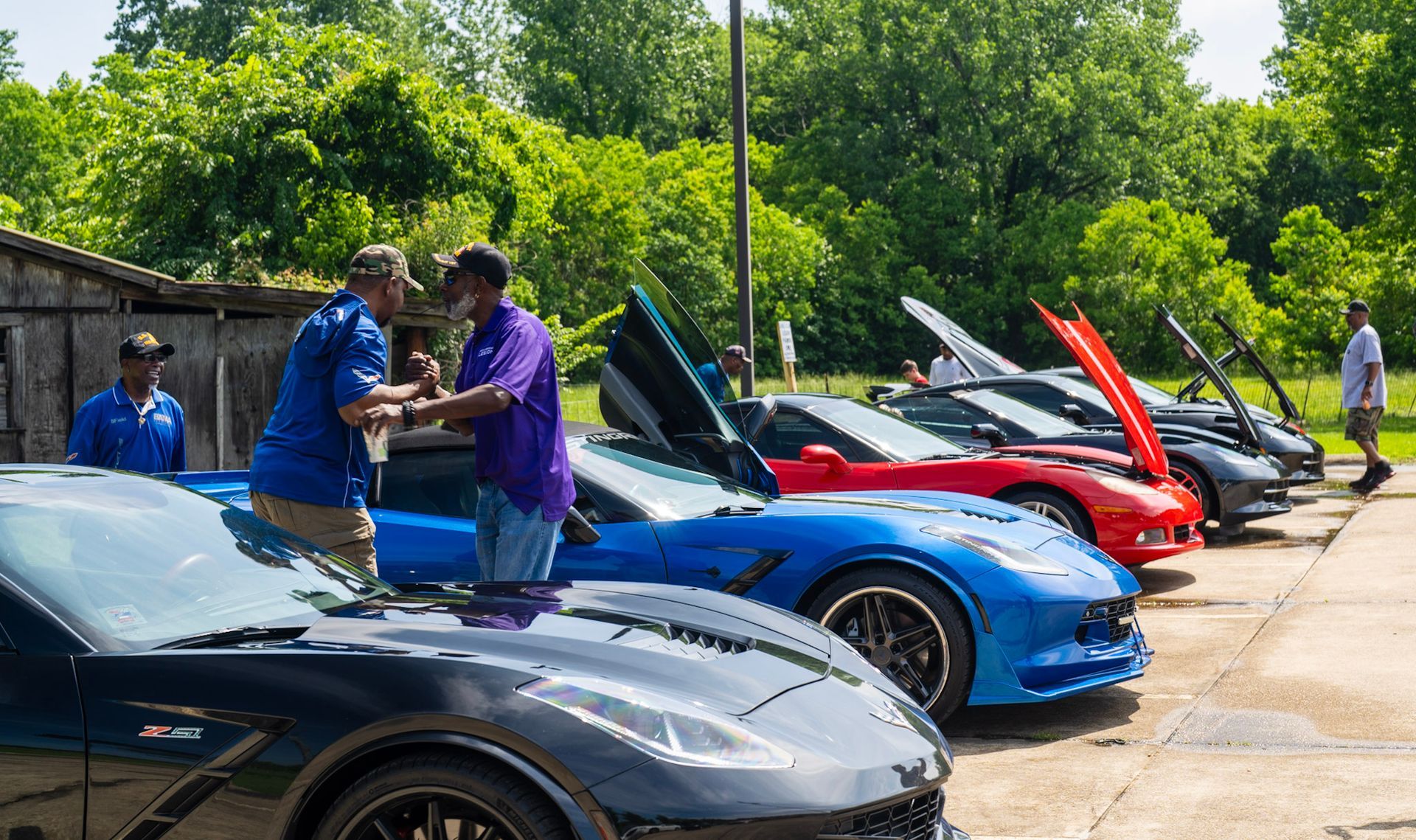 Two people shake hands at an outdoor car show featuring several parked sports cars with hoods or doors open.