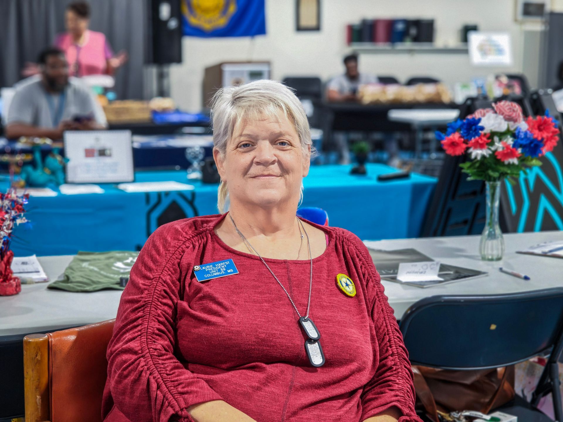 A person in a red shirt sits at a table in a meeting space, with a bouquet of red, white, and blue flowers nearby.