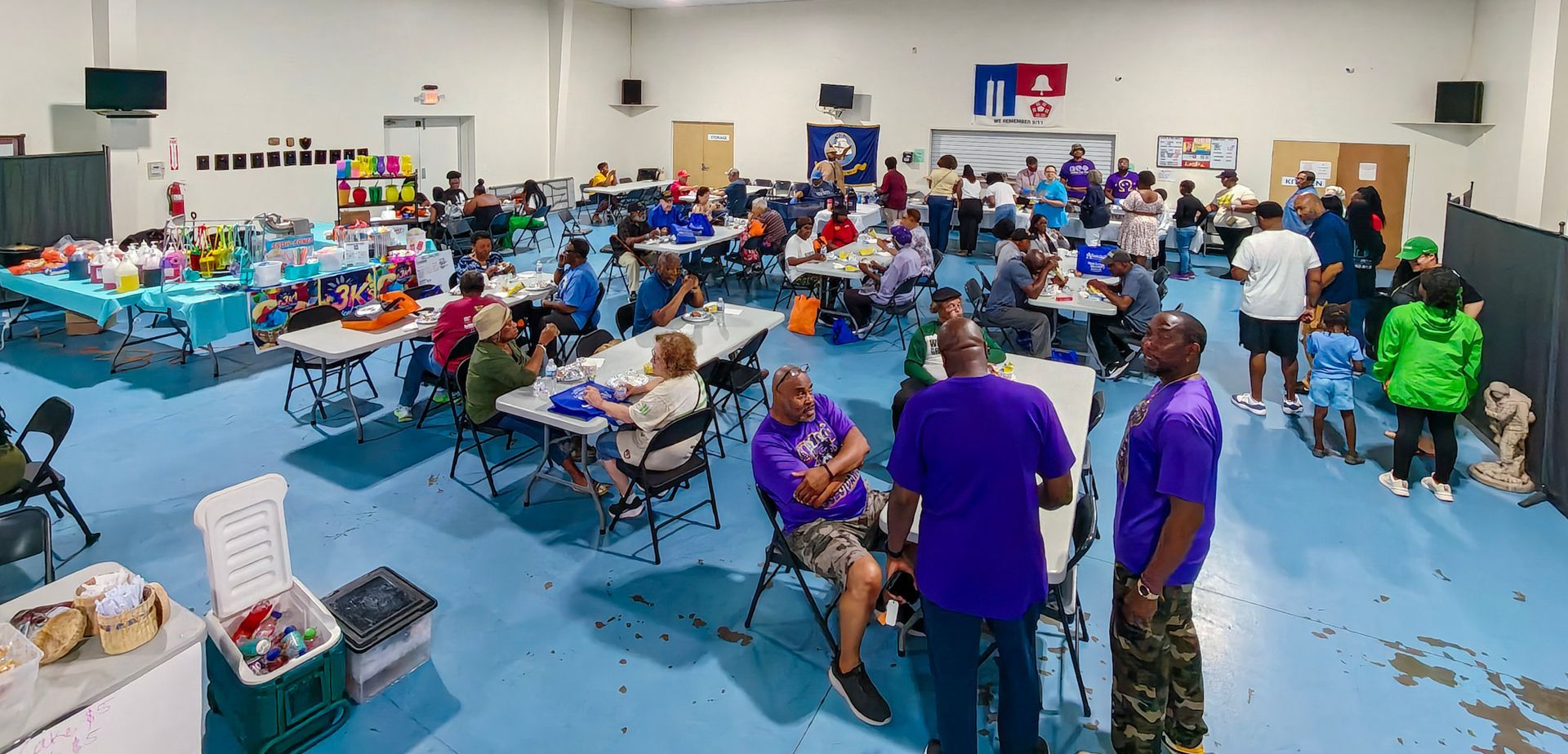 A large, brightly lit indoor space with people sitting and standing around tables at a community or group gathering.