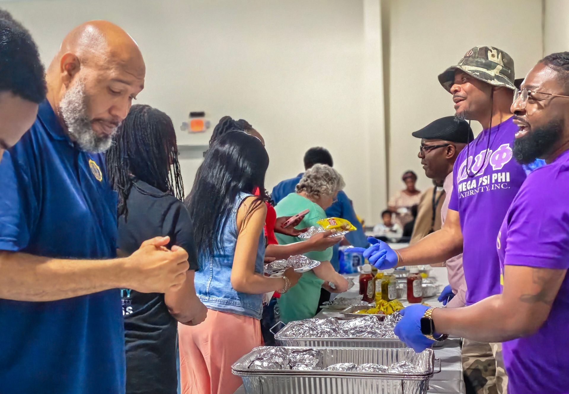 People in a bright room serving prepared meals from foil-covered pans to others in a line.