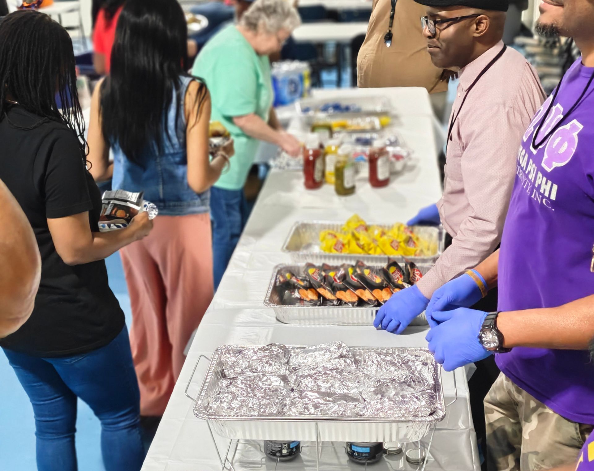 People serve food in foil trays at a catered event with a long table of snacks and condiments.