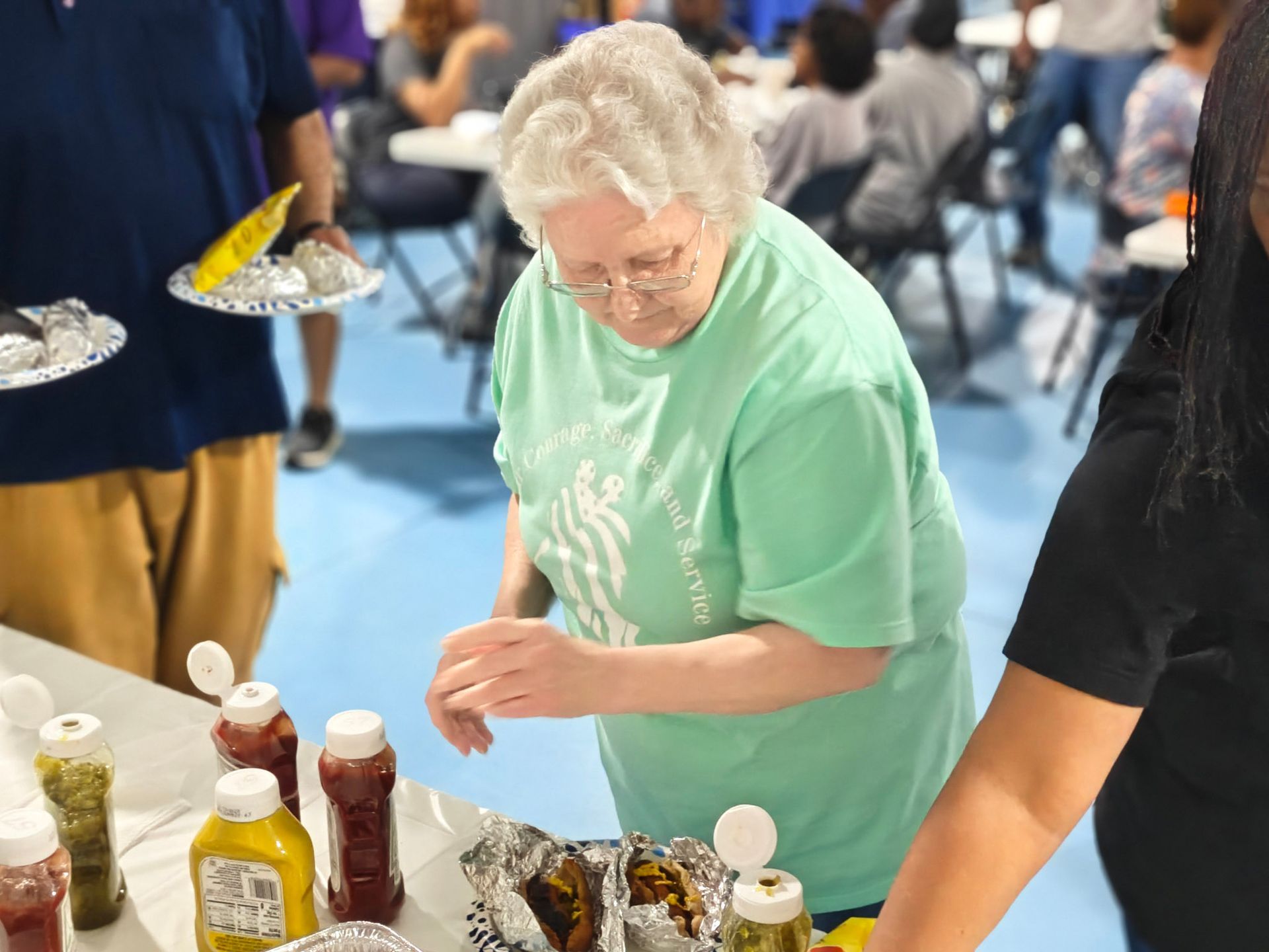 A person in a light green shirt prepares food at a table with condiment bottles and foil-wrapped items.