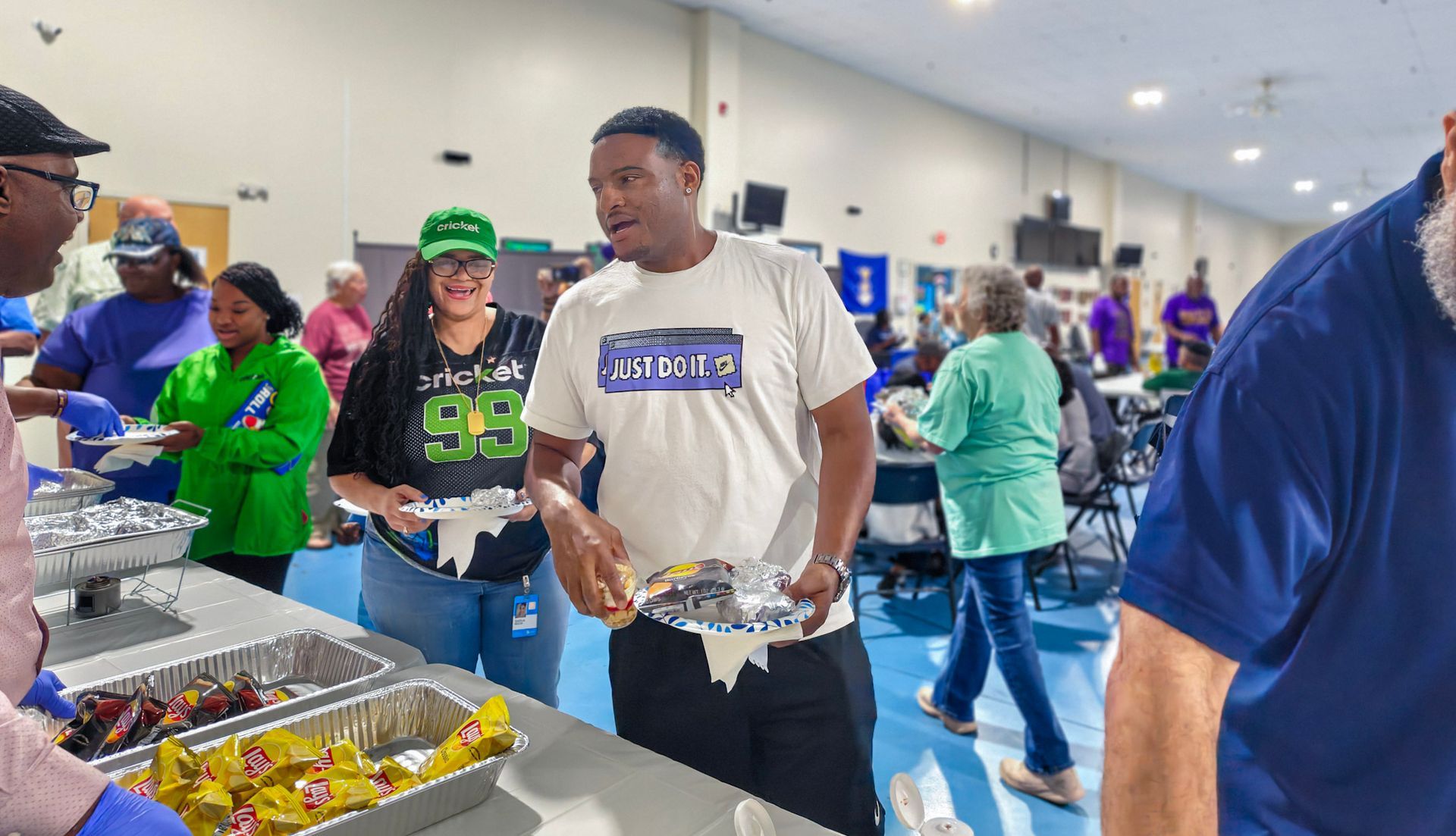 People serve food from buffet trays in a brightly lit, spacious community center or gymnasium setting.