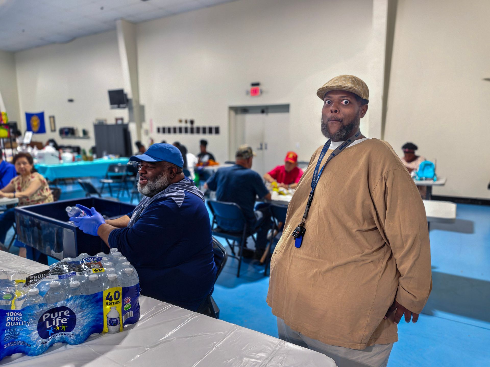 A person in a tan shirt and cap stands in an activity room, while another person prepares water bottles in the background.