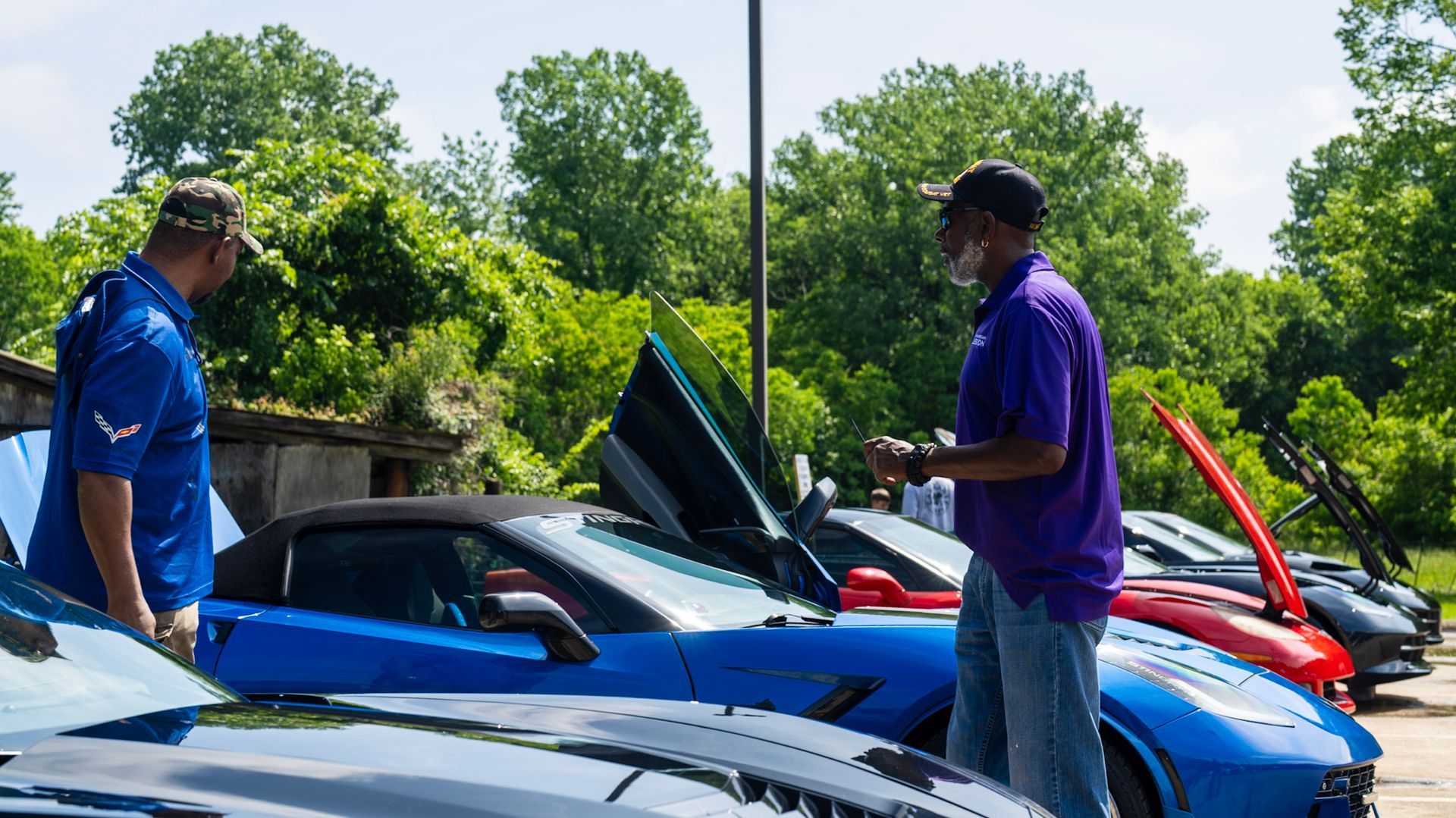 Two people stand near a bright blue sports car with its door open at an outdoor car show on a sunny day.