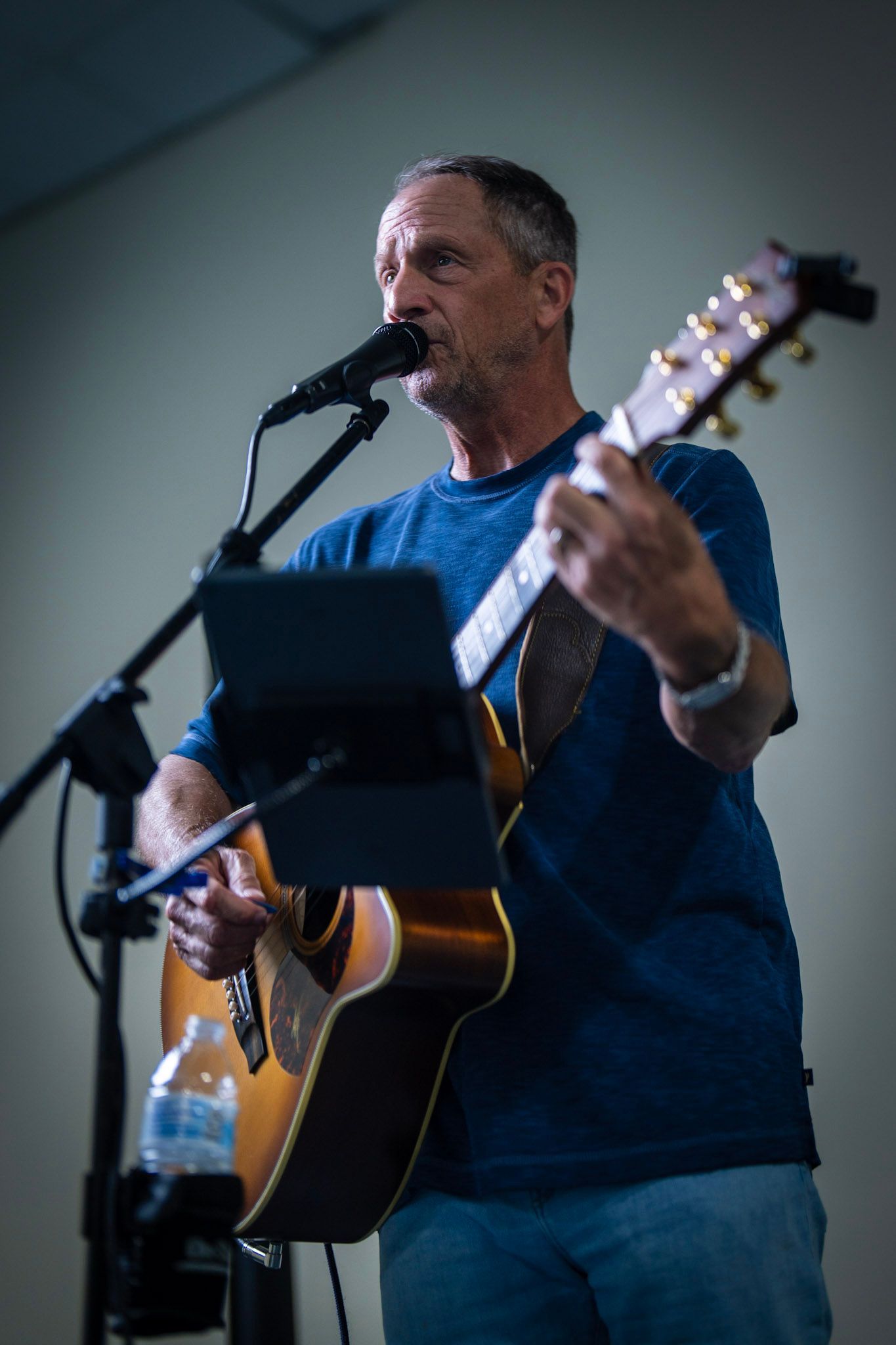 A performer in a blue t-shirt plays an acoustic guitar and sings into a microphone, with a music stand and water bottle.